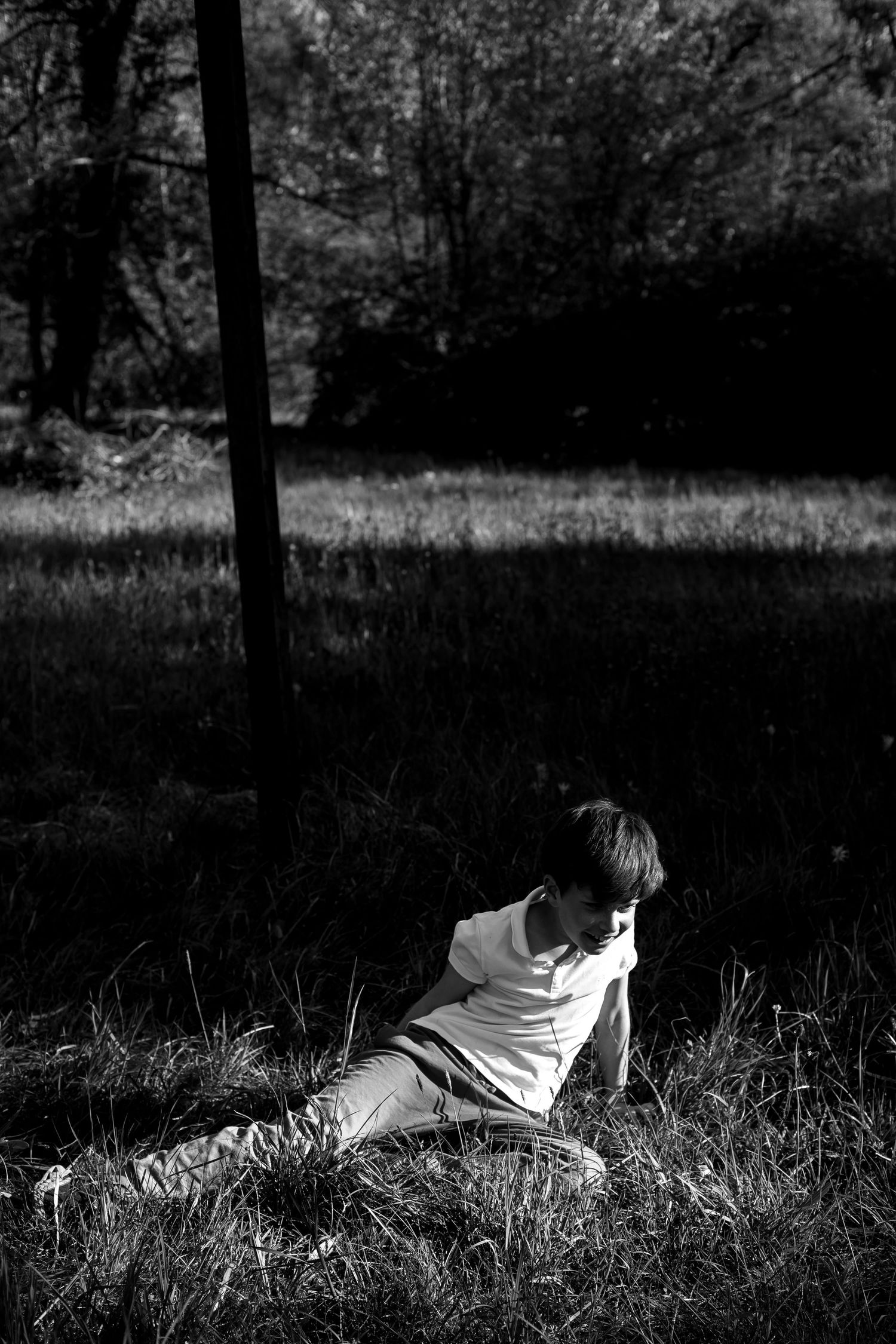 Black and white photo of a boy sitting alone in sunlit grass field near a dark pole.