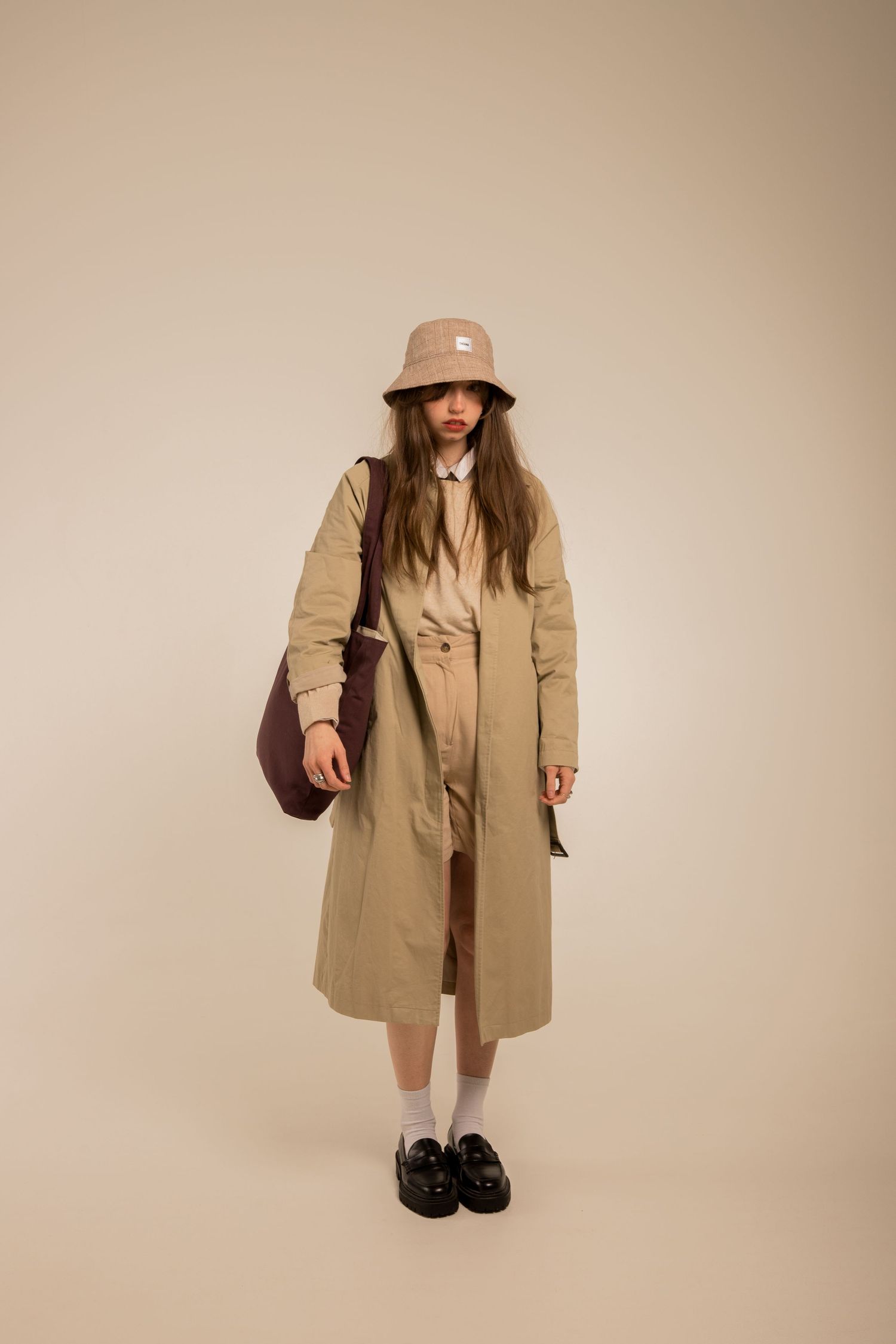 Woman in beige trench coat and bucket hat carrying burgundy bag on neutral background.