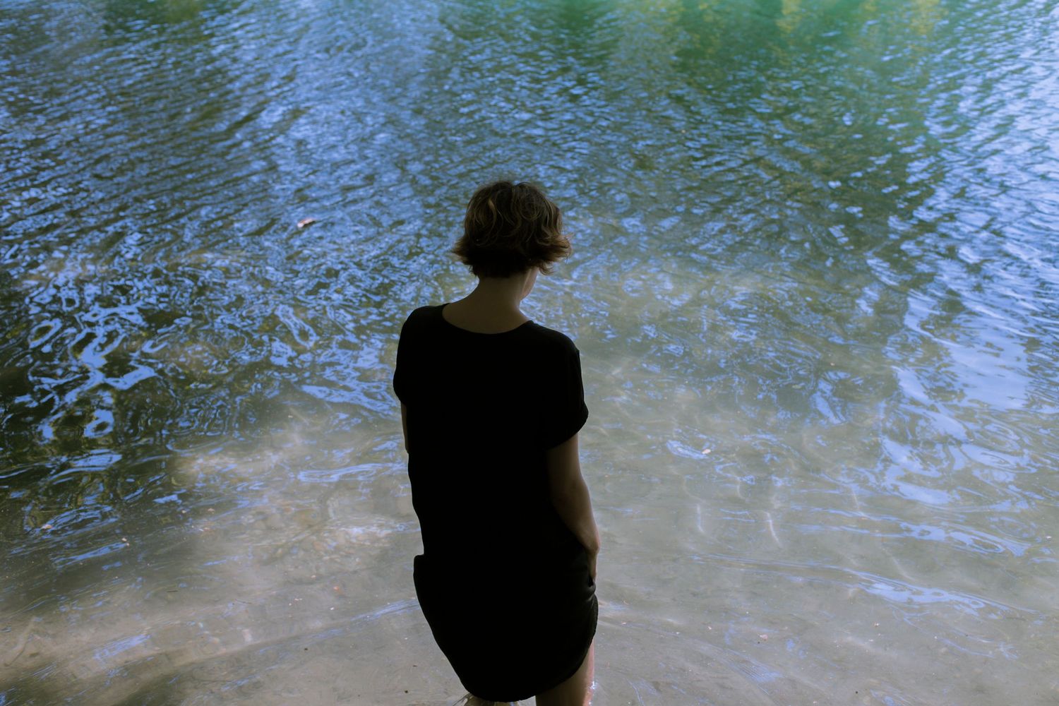 Person in black dress standing at the edge of a calm reflective body of water from behind.
