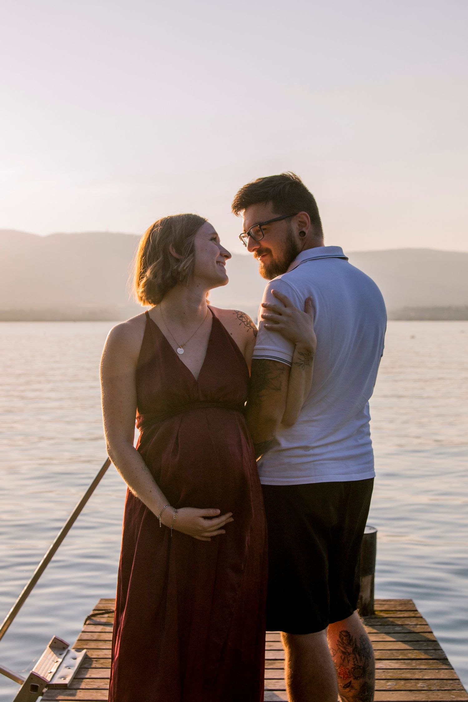 Expecting couple embracing on wooden dock at golden sunset, smiling at each other over calm lake.