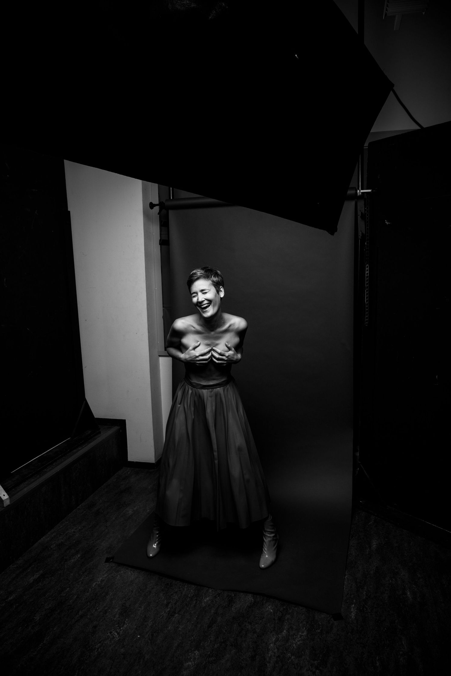 Black and white behind-the-scenes studio photo of woman in tulle skirt covering chest laughing.