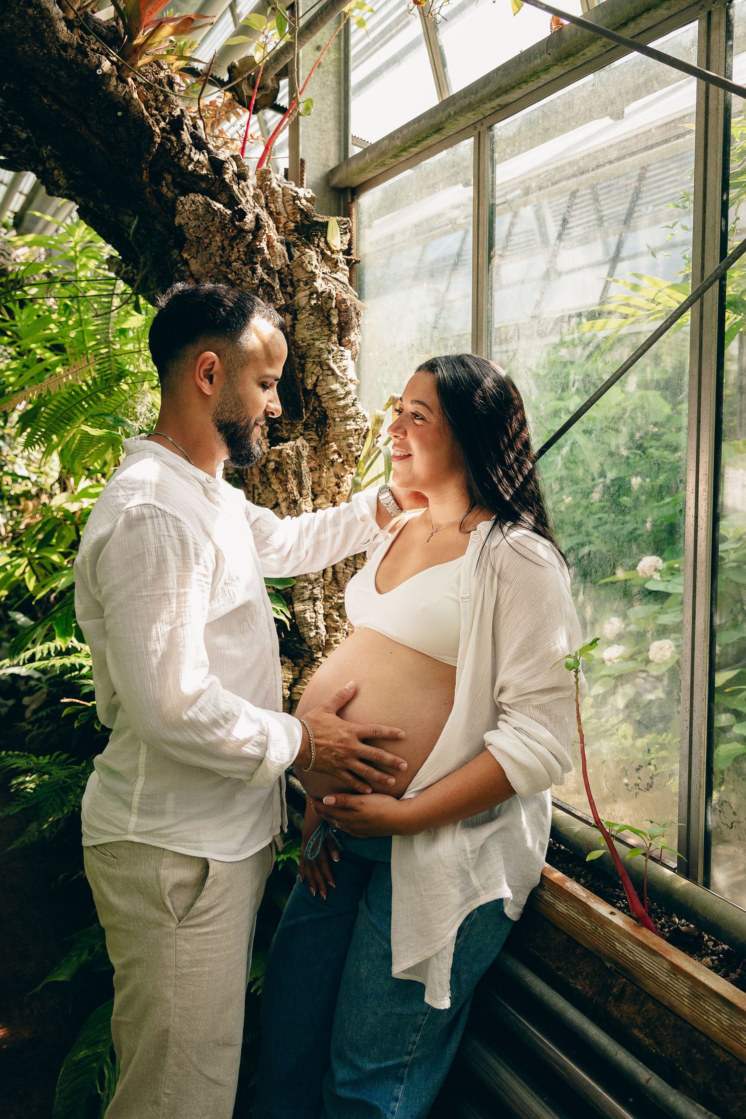 Pregnant couple in white outfits share a tender moment in a lush greenhouse, man gently touching woman's bare belly.
