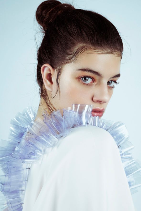 Young woman with hair bun wearing avant-garde white outfit with translucent ruffled collar against light blue background.