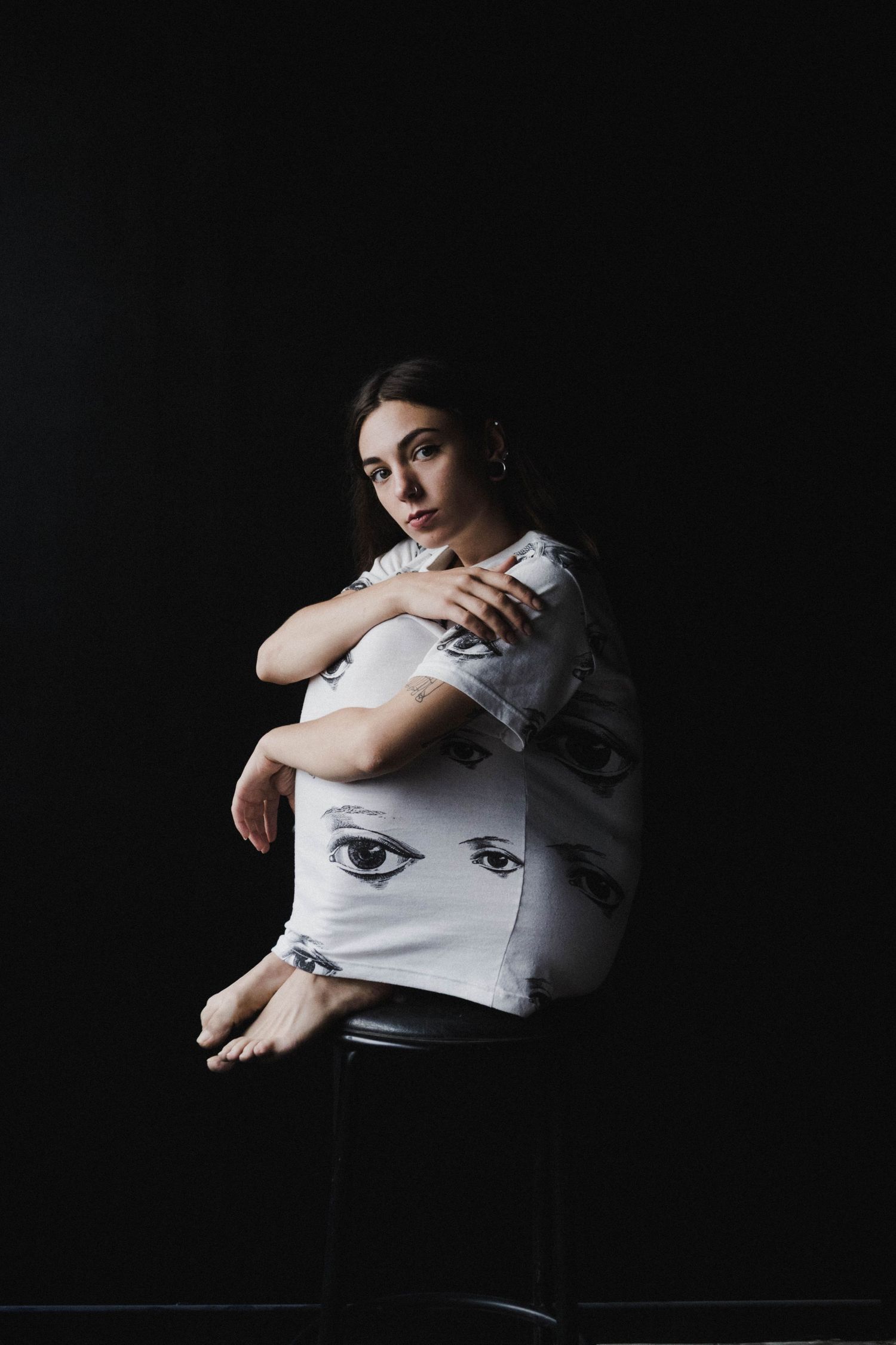 Young woman in printed eye-pattern white shirt seated on stool with arms crossed against black background.