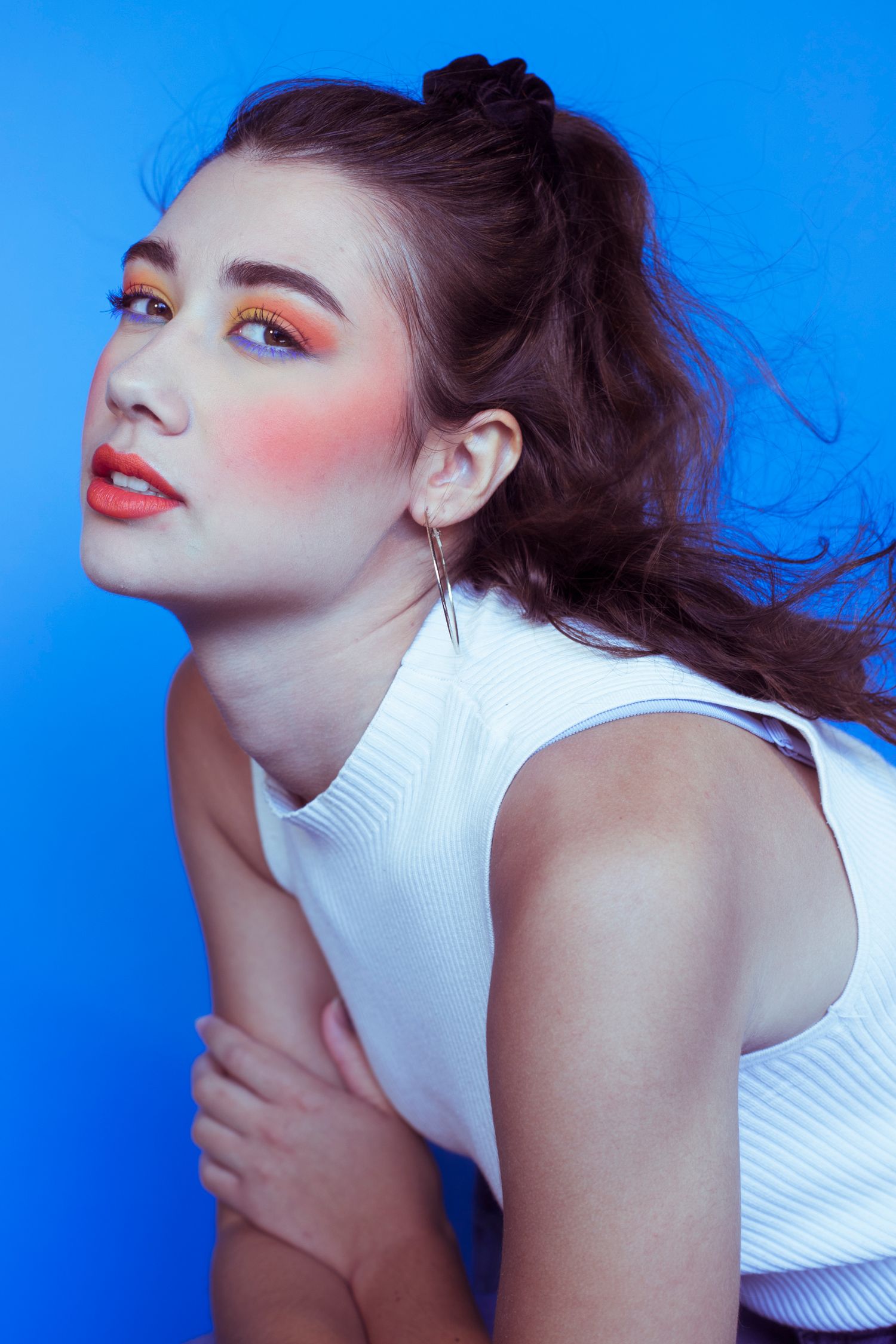 Brunette model with bold orange eyeshadow in white sleeveless top leans forward against bright blue background.