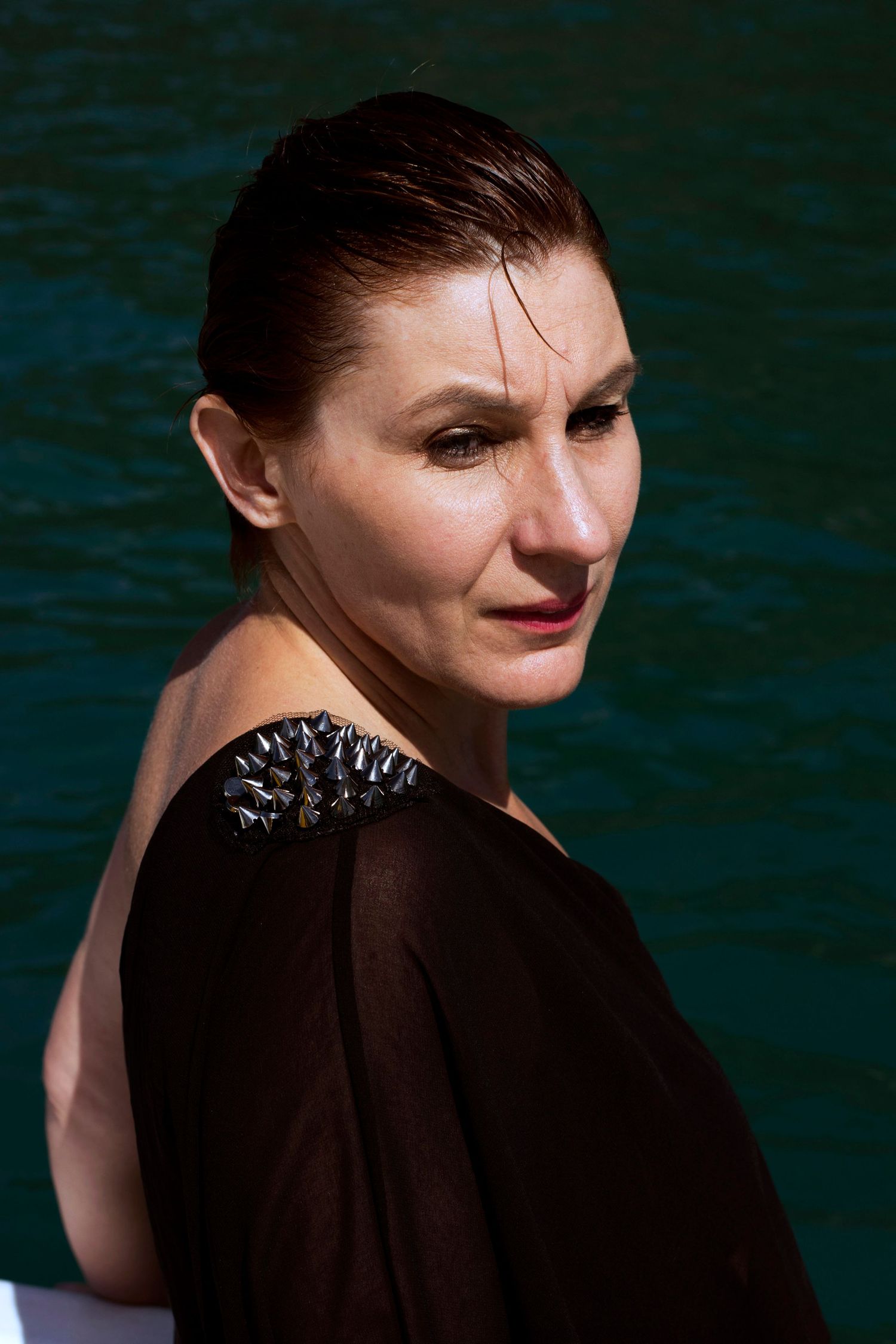 Close-up portrait of a woman in black off-shoulder top with spiked detail near turquoise water.