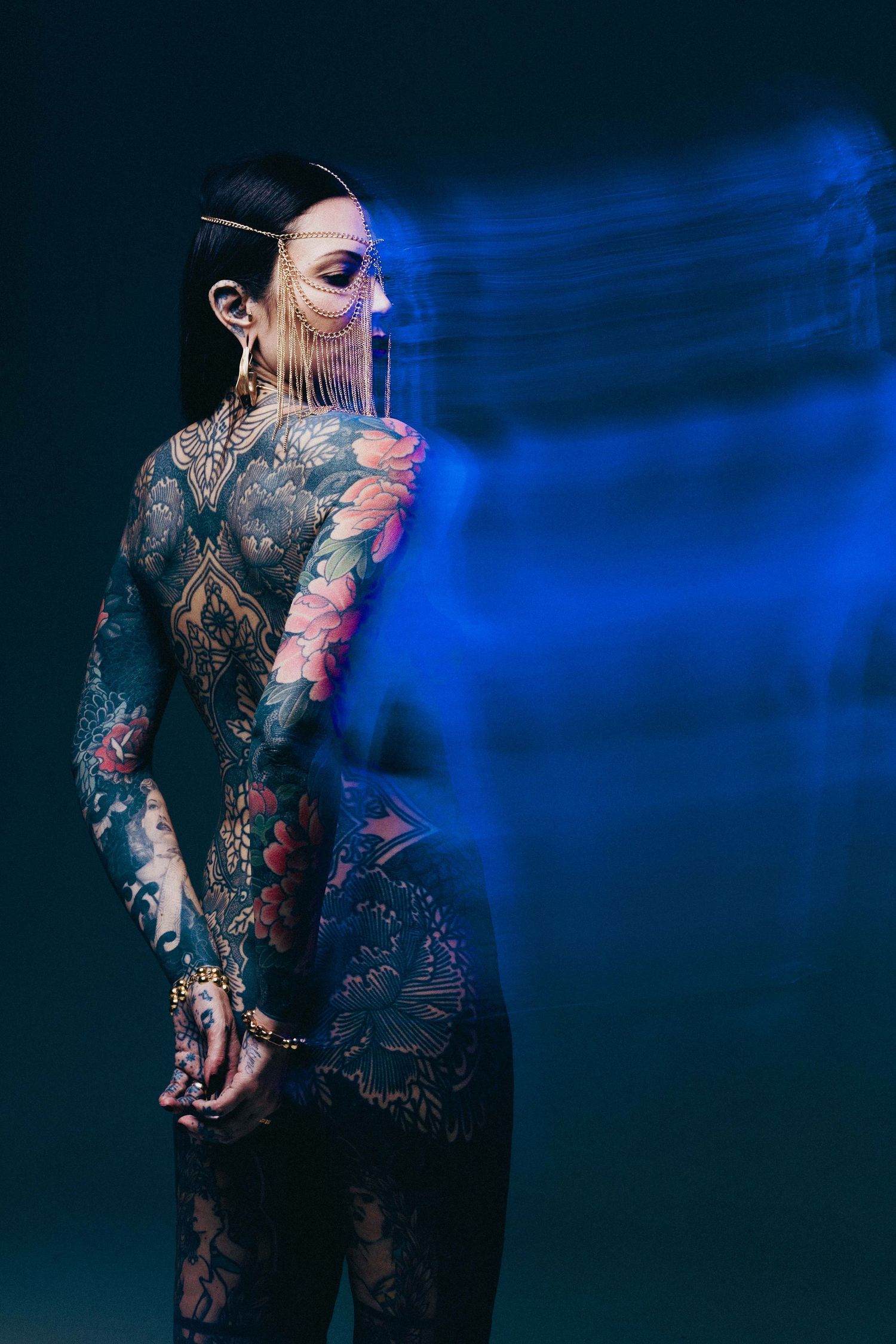 Back view of tattooed woman with full body ink wearing gold headpiece in blue studio light.