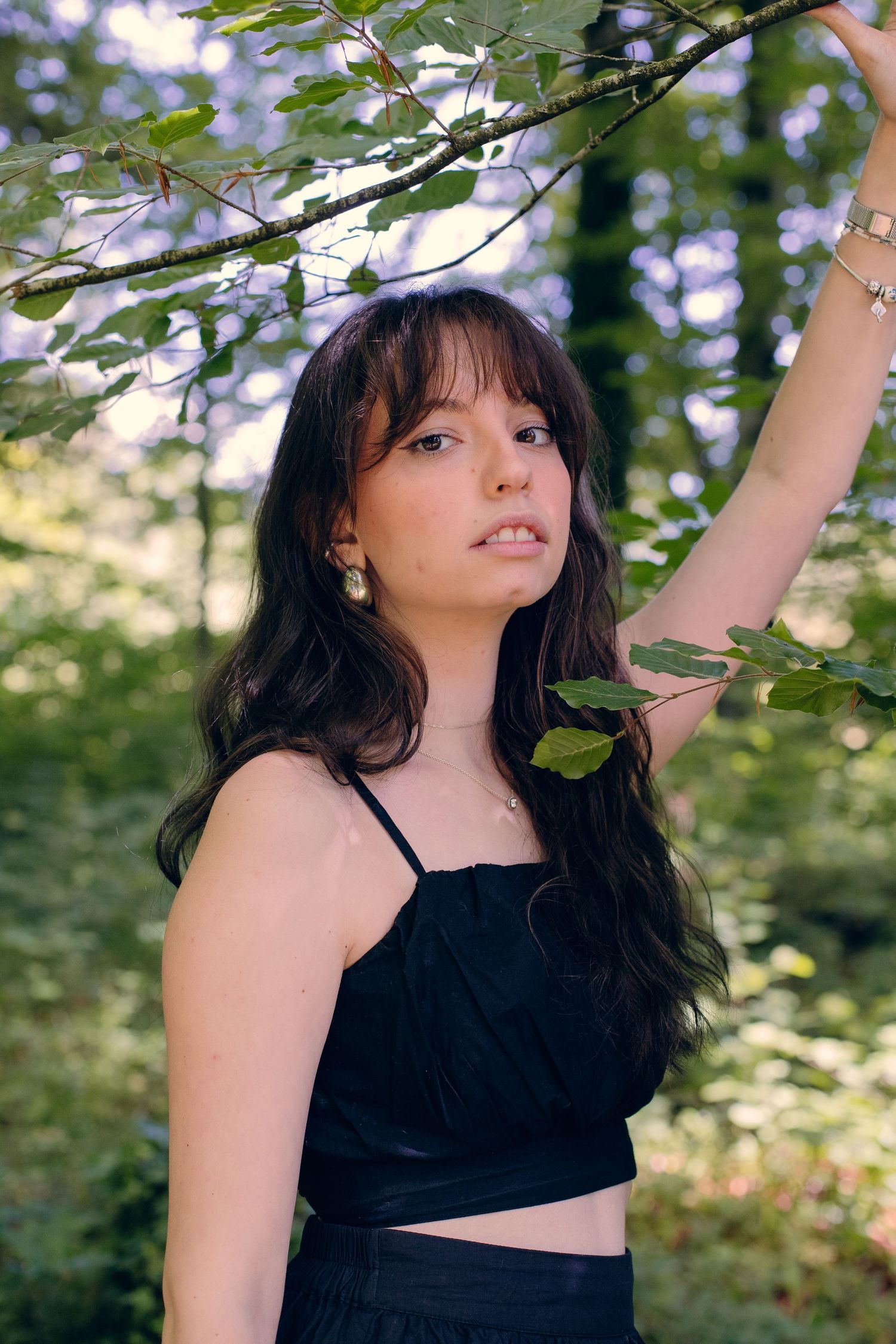Young woman in black crop top reaching up to touch leafy tree branch outdoors.
