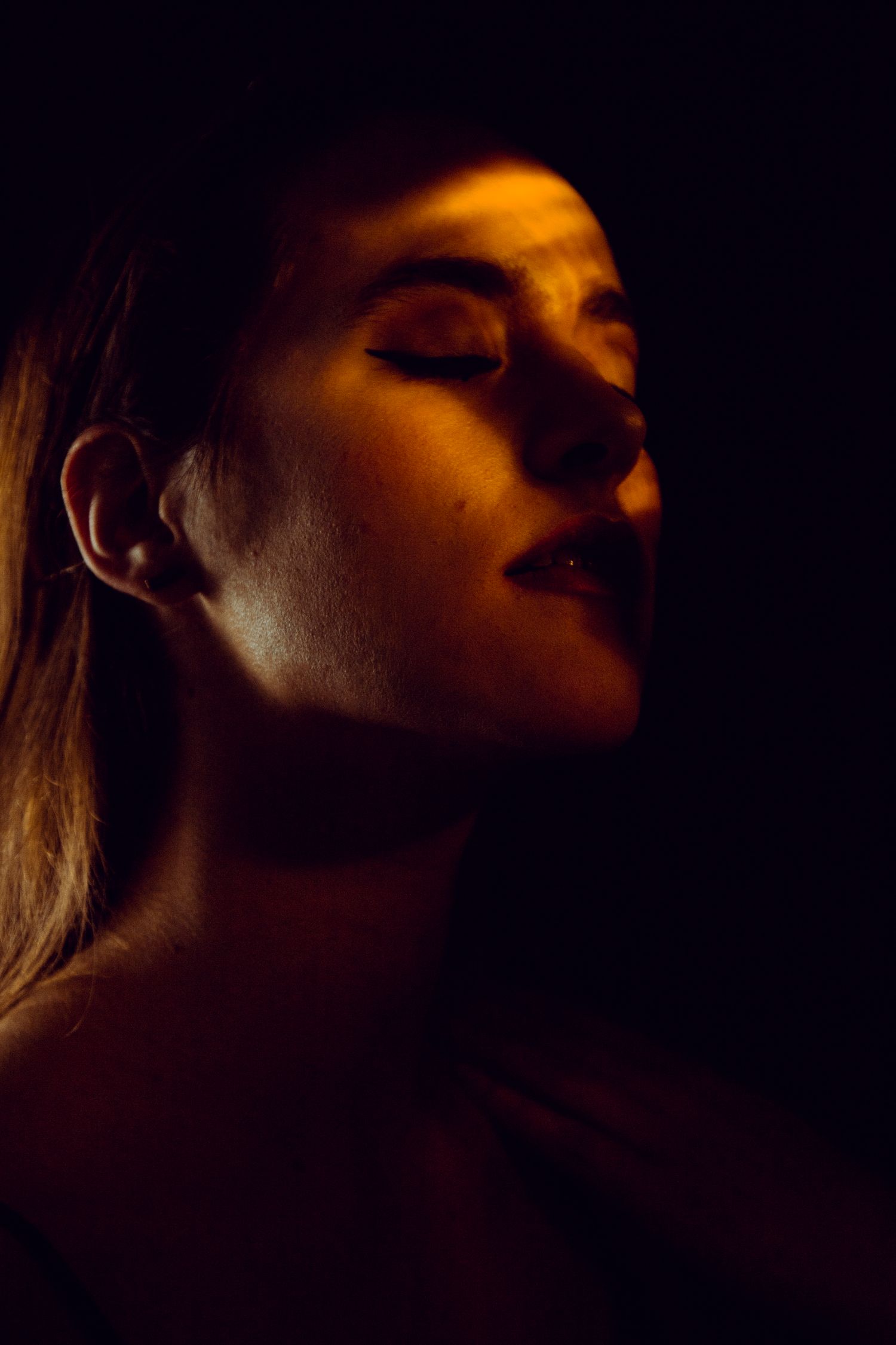 Warm amber low-key portrait of a woman with closed eyes and dramatic side lighting on dark background.