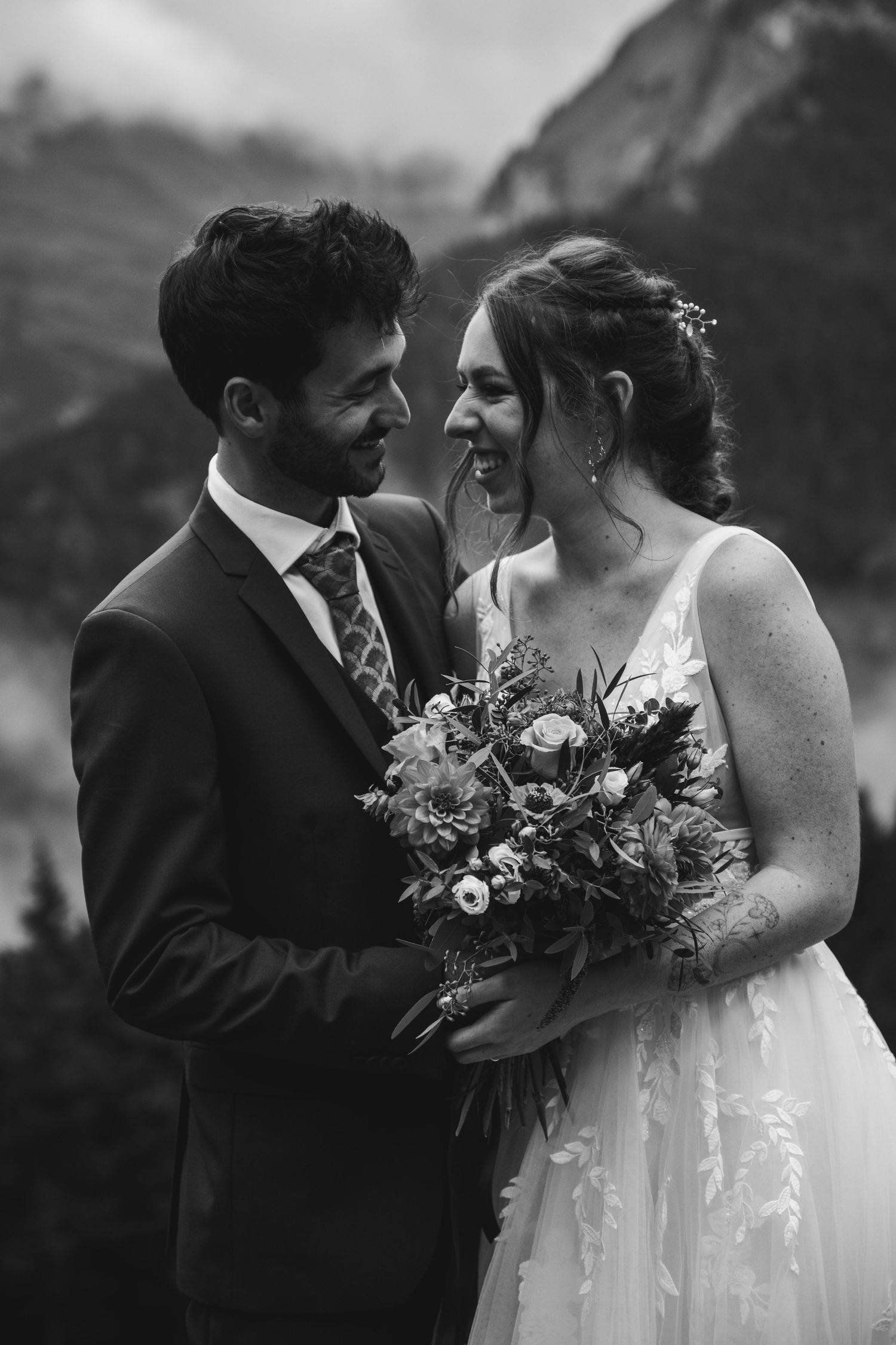 Black and white photo of laughing bride and groom embracing with mountain backdrop.