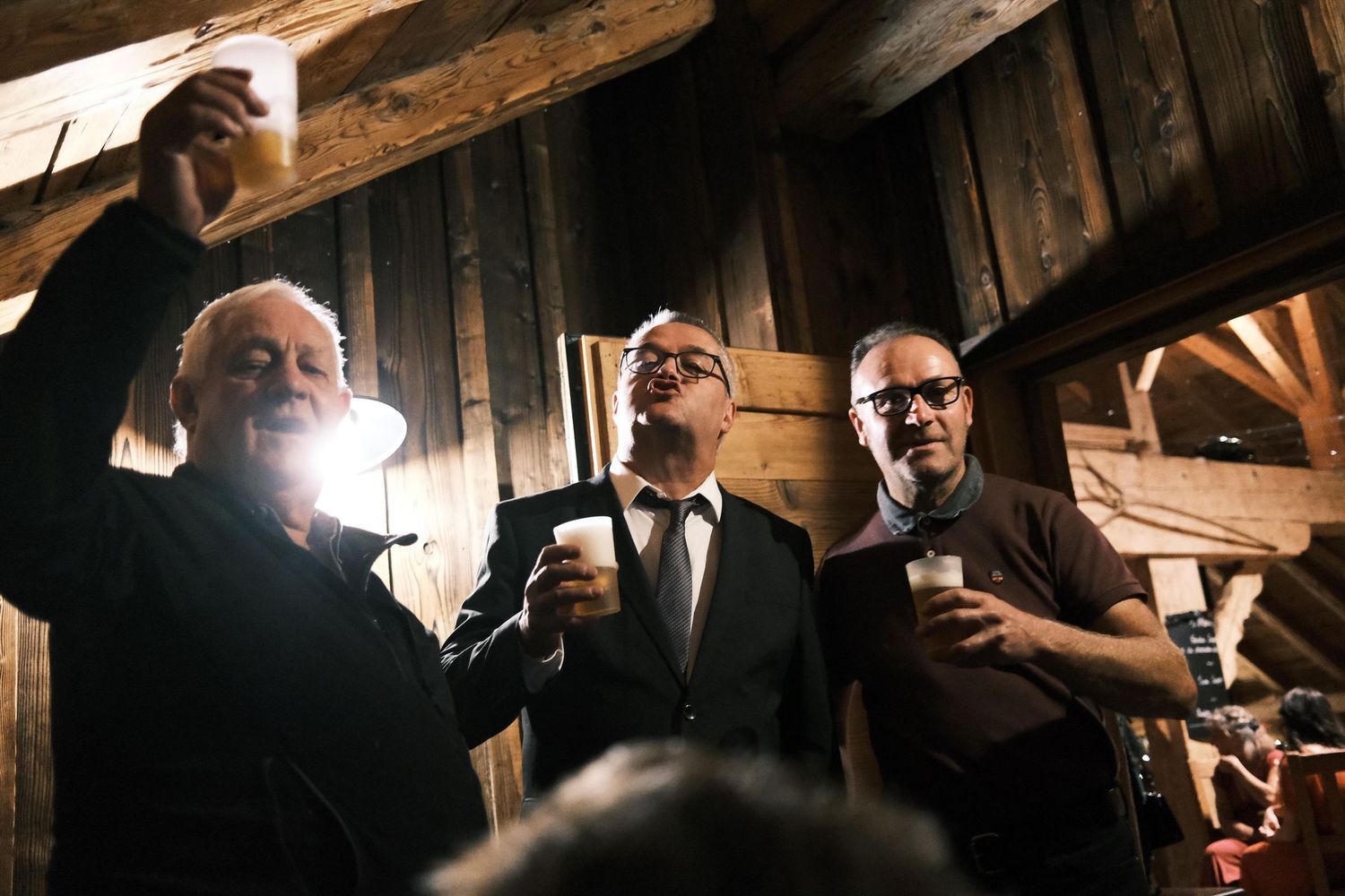 Three men in formal attire toasting with drinks inside a rustic wooden barn at a wedding reception.