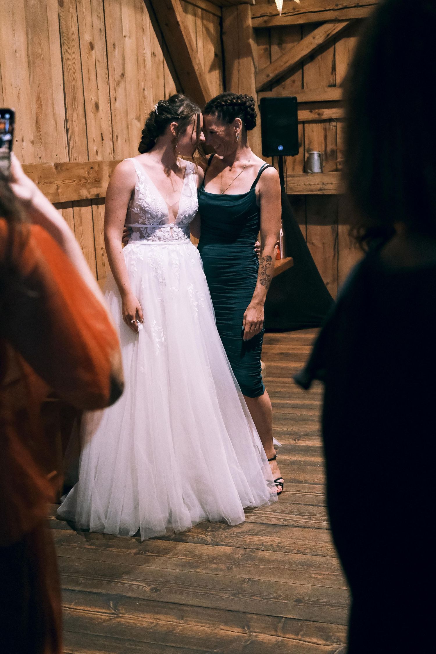 Bride in white tulle gown shares tender moment with woman in green dress at barn wedding.