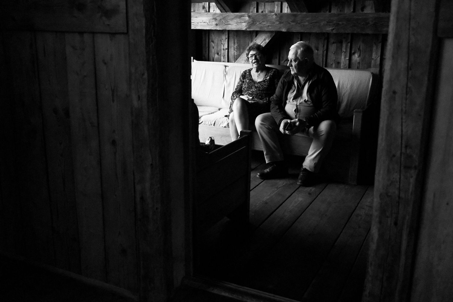 Black and white photo of elderly couple sitting together in rustic wooden barn doorway.