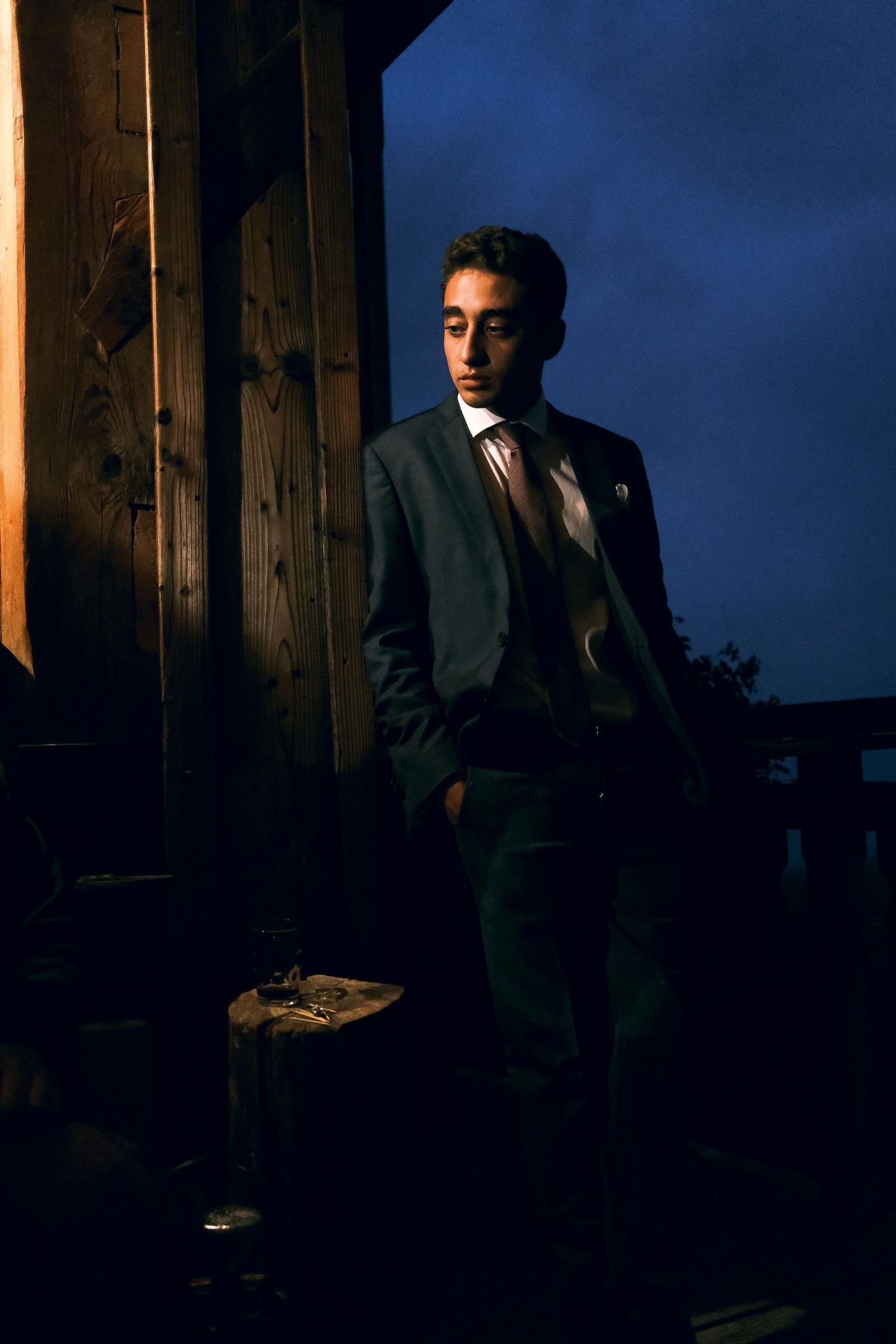 Elegant young man in dark suit poses dramatically against wooden pillar under a deep blue night sky.