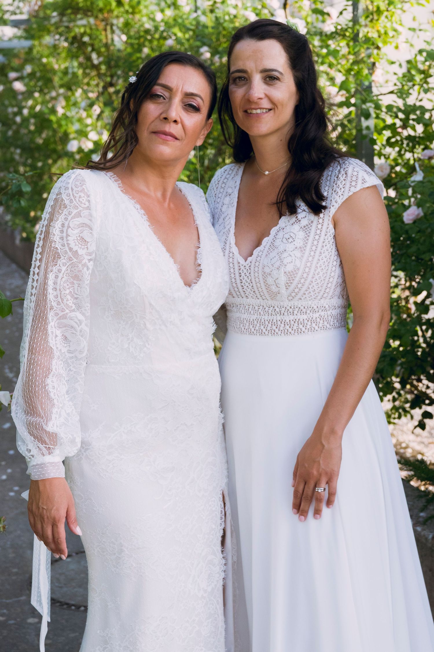 Two brides in white wedding dresses pose together for a portrait in a garden setting.