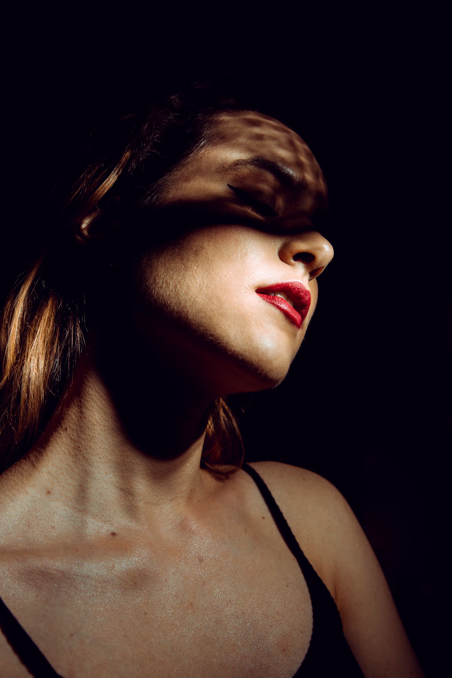 Woman with red lips and shadow across her eyes posed against dark background in dramatic portrait photography.