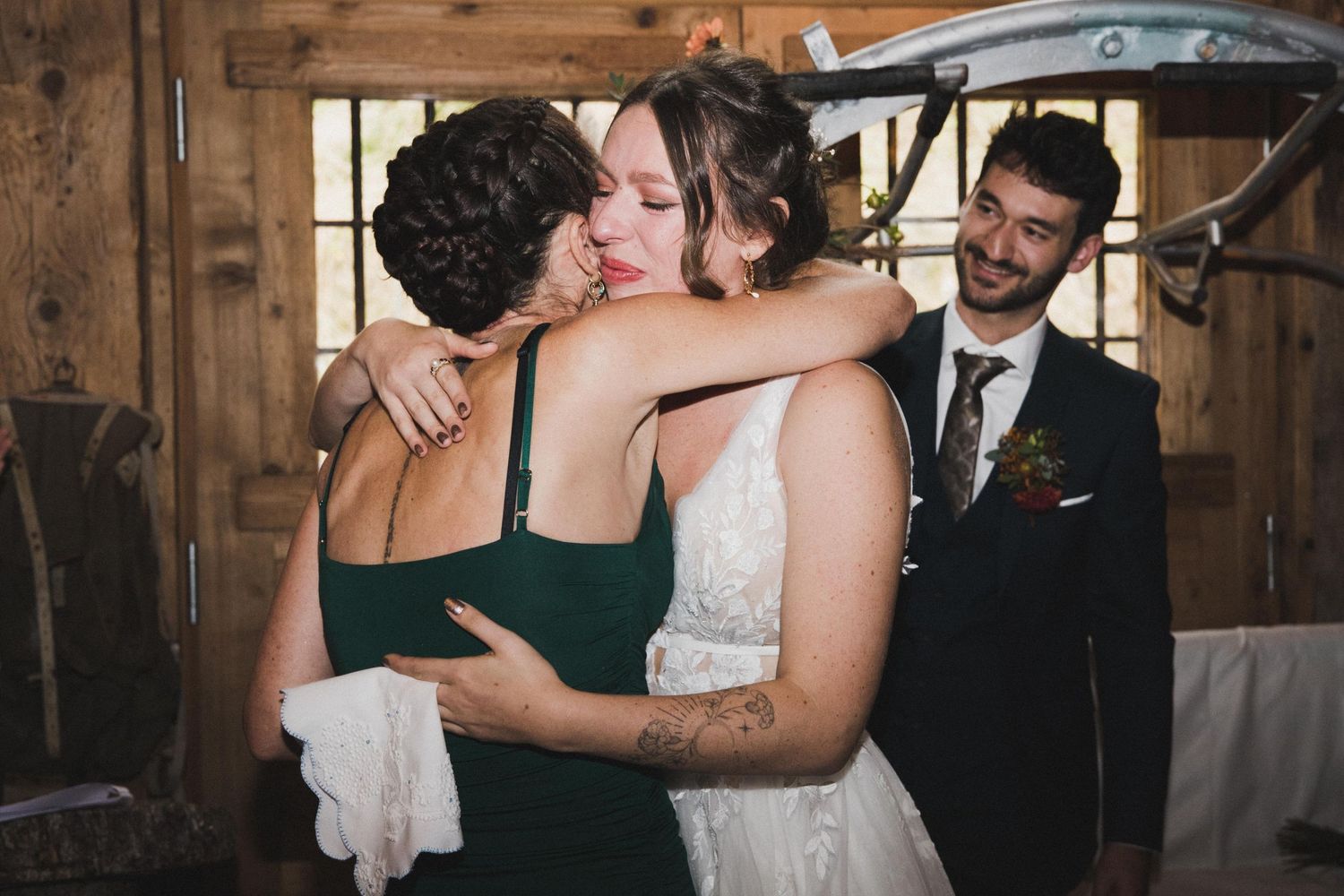 Bride in white lace gown embracing bridesmaid in green dress as groom smiles in background.