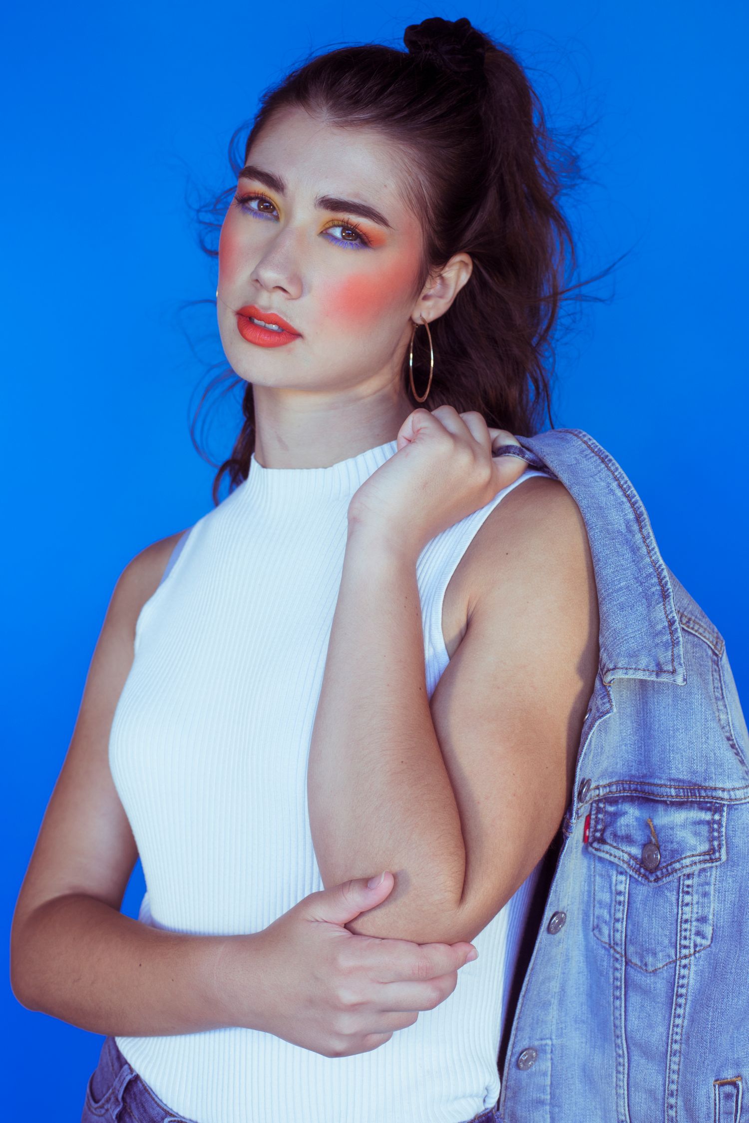 Model in white sleeveless top holding denim jacket with vibrant orange makeup against blue background.