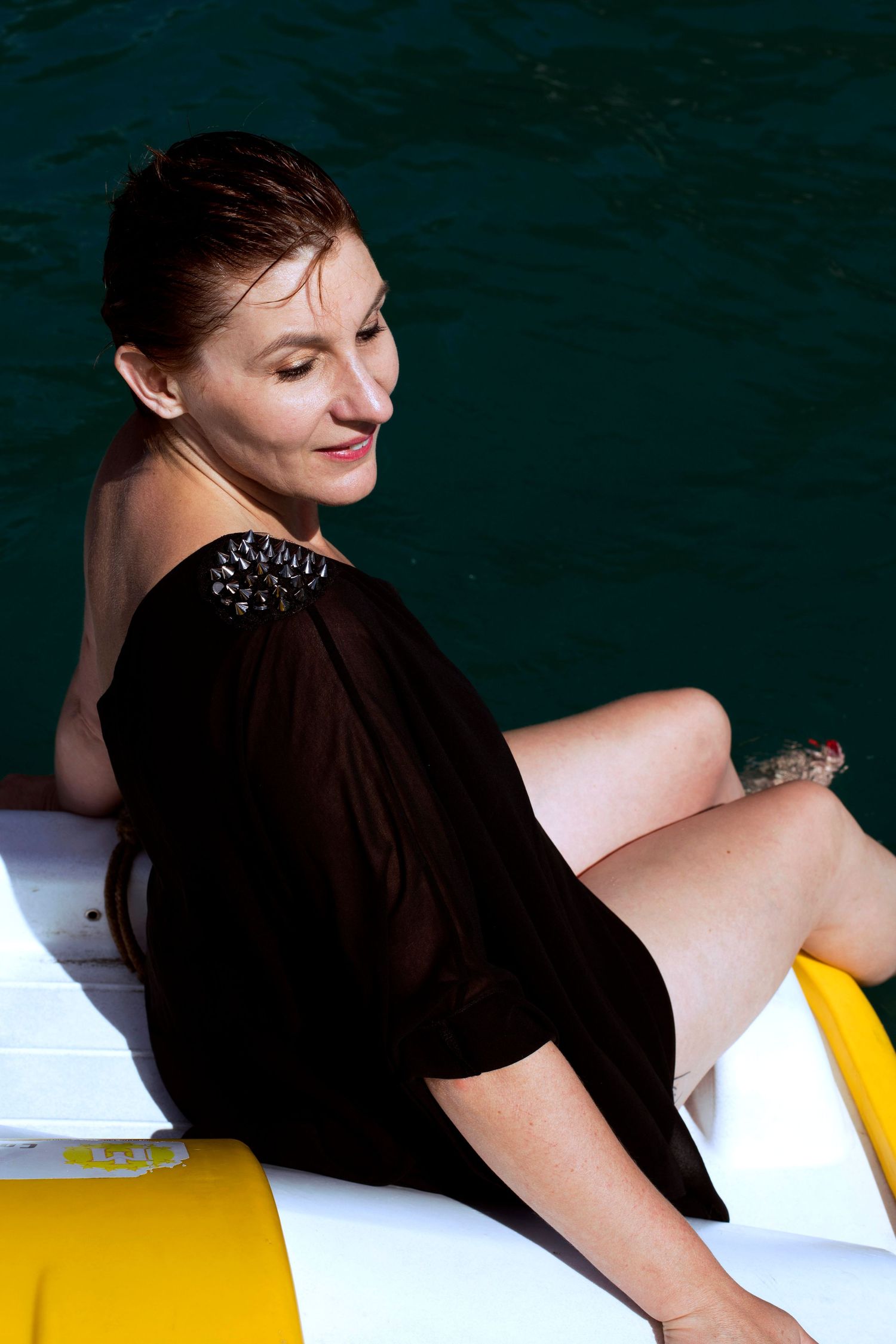 Woman in black sheer off-shoulder top relaxing on a yellow boat over deep turquoise water.