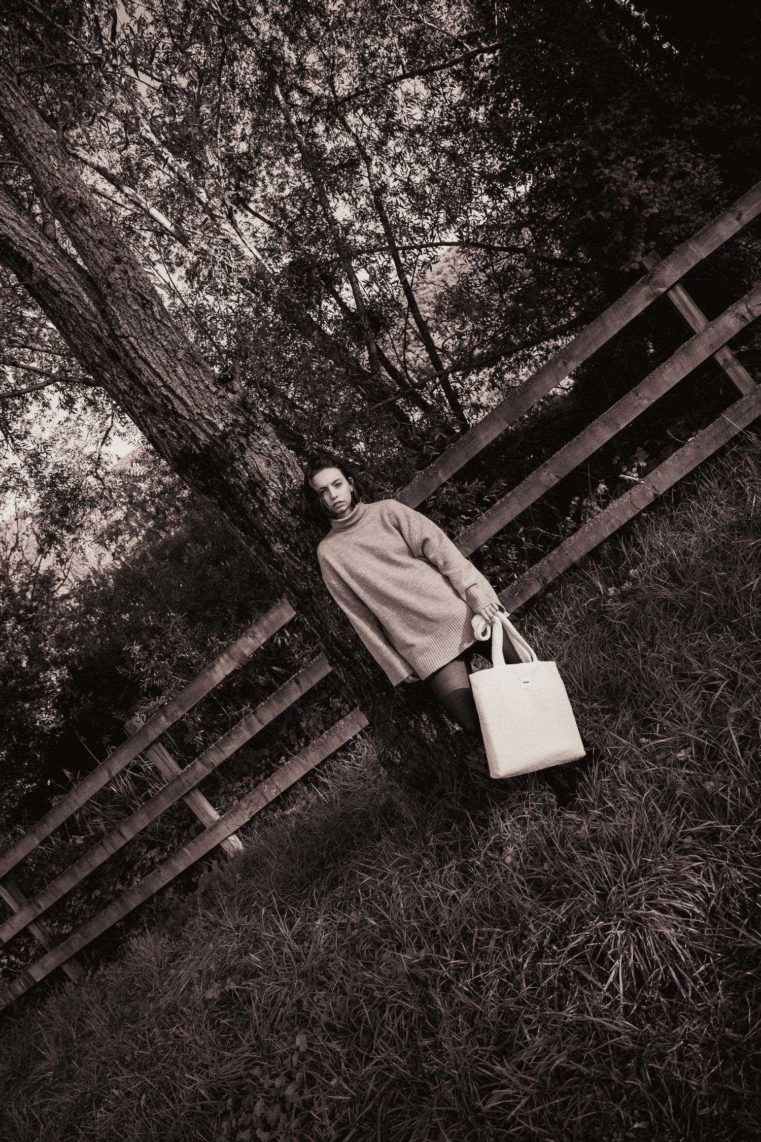 Black and white photo of a woman in a knit sweater holding a tote bag by a wooden fence outdoors.