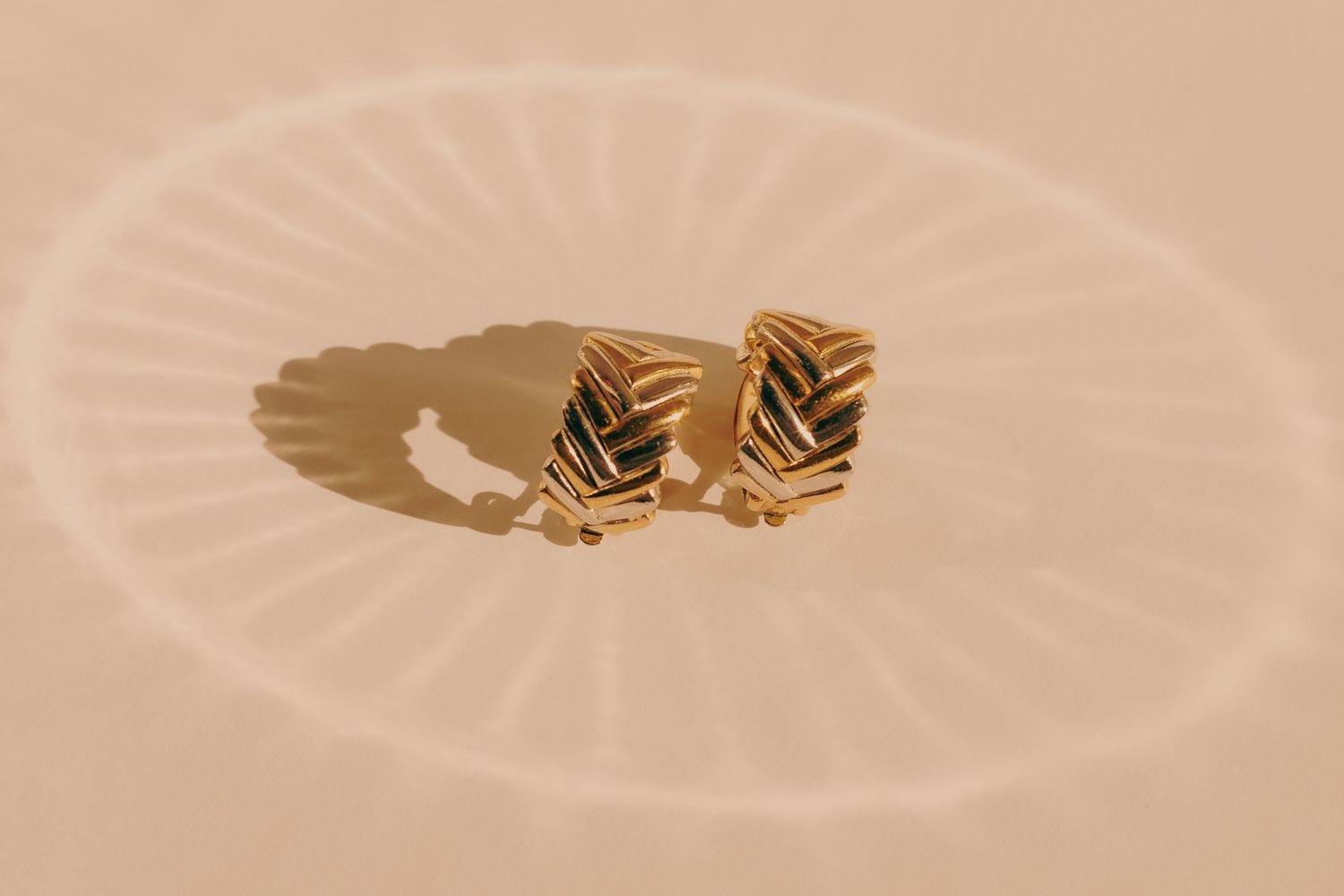 Two gold chevron textured clip-on earrings casting shadows on a warm beige surface.