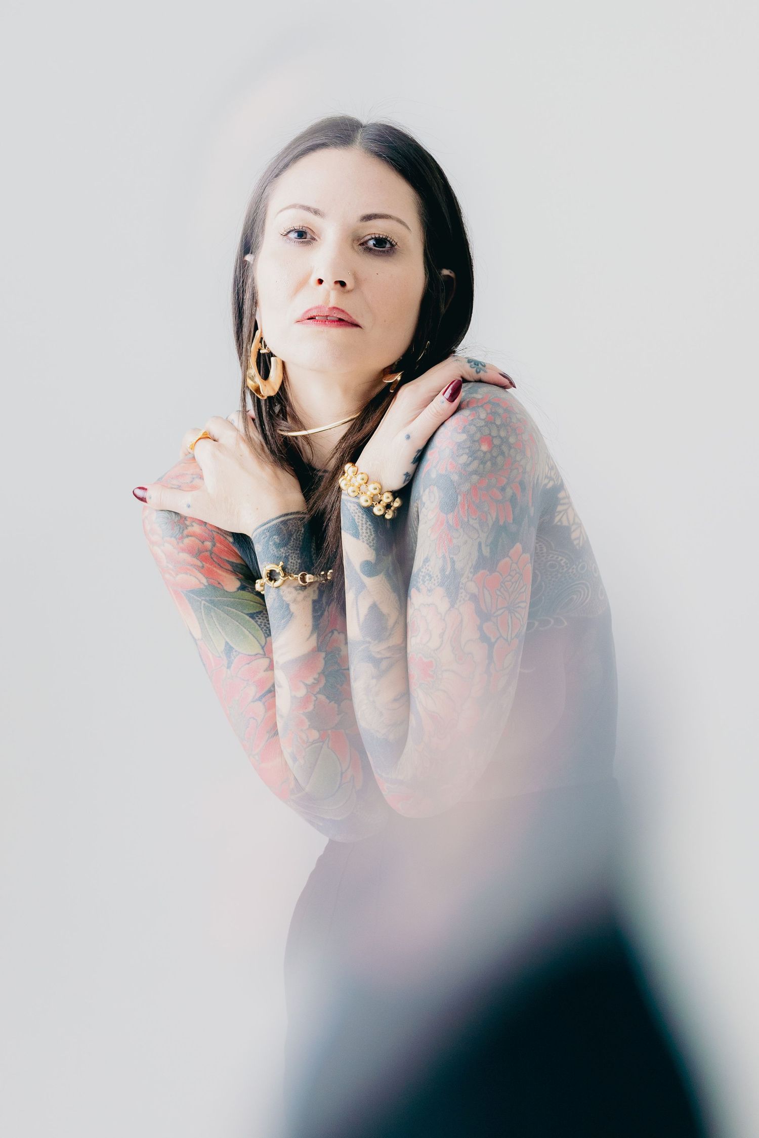 Tattooed woman with gold jewelry crosses her arms over her chest in a bright studio portrait.