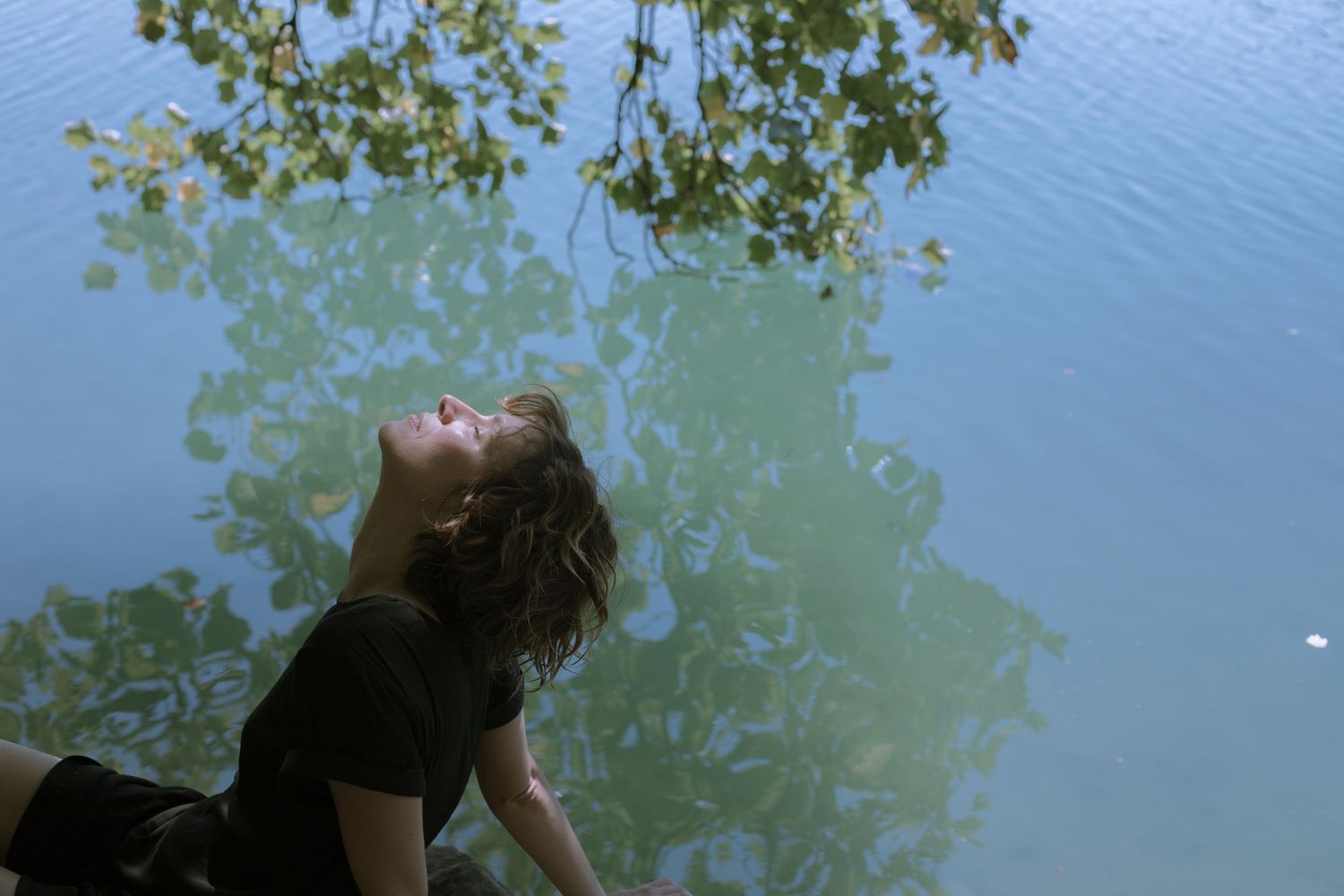 Woman leaning back with eyes closed near a tranquil lake surrounded by green foliage.