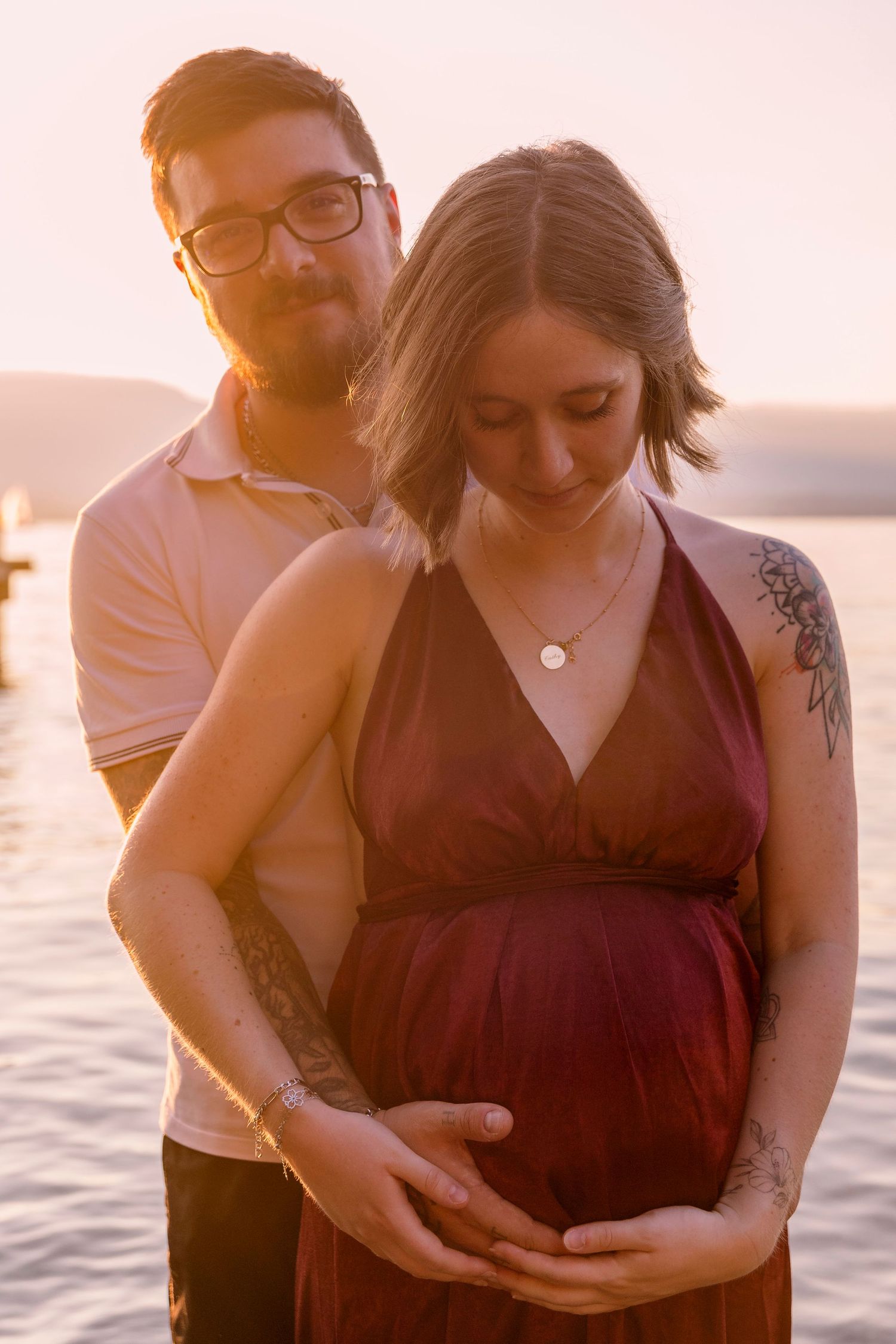 Man embracing pregnant partner from behind at sunset, both cradling her baby bump over serene lake.