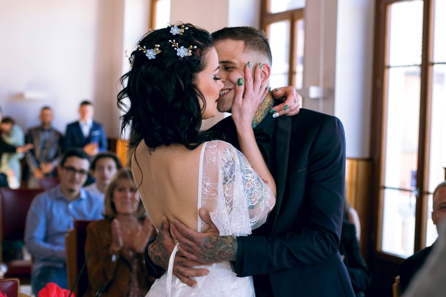 Bride and groom share first kiss during indoor wedding ceremony while guests applaud in background.