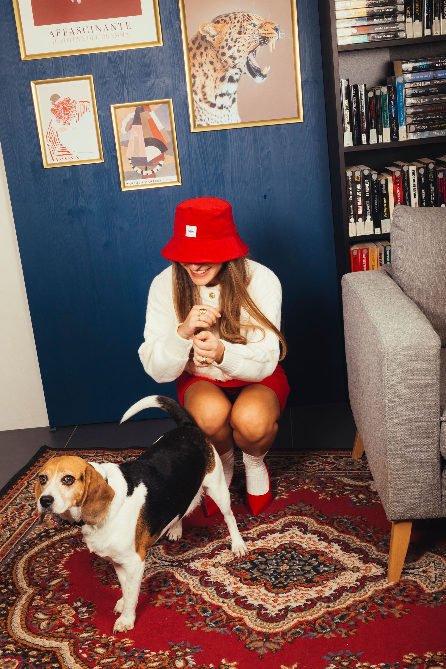 Woman in red bucket hat and white sweater crouches to pet a beagle dog on a colorful rug indoors.