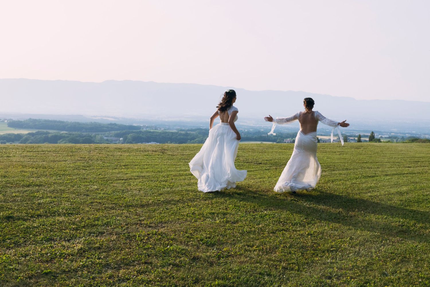 Two brides in white dresses run joyfully across a green field with scenic mountain views.