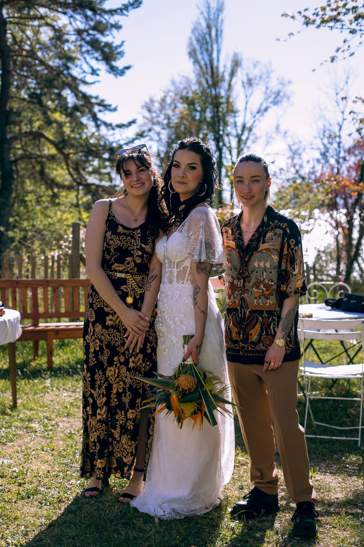 Bride in white lace dress holding tropical bouquet posing with two guests in patterned outfits outdoors.