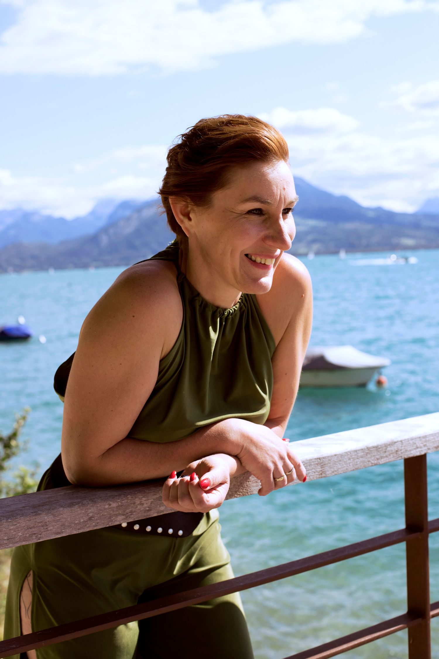 Smiling redhead woman in olive green dress leaning on a railing with mountain lake view behind her.