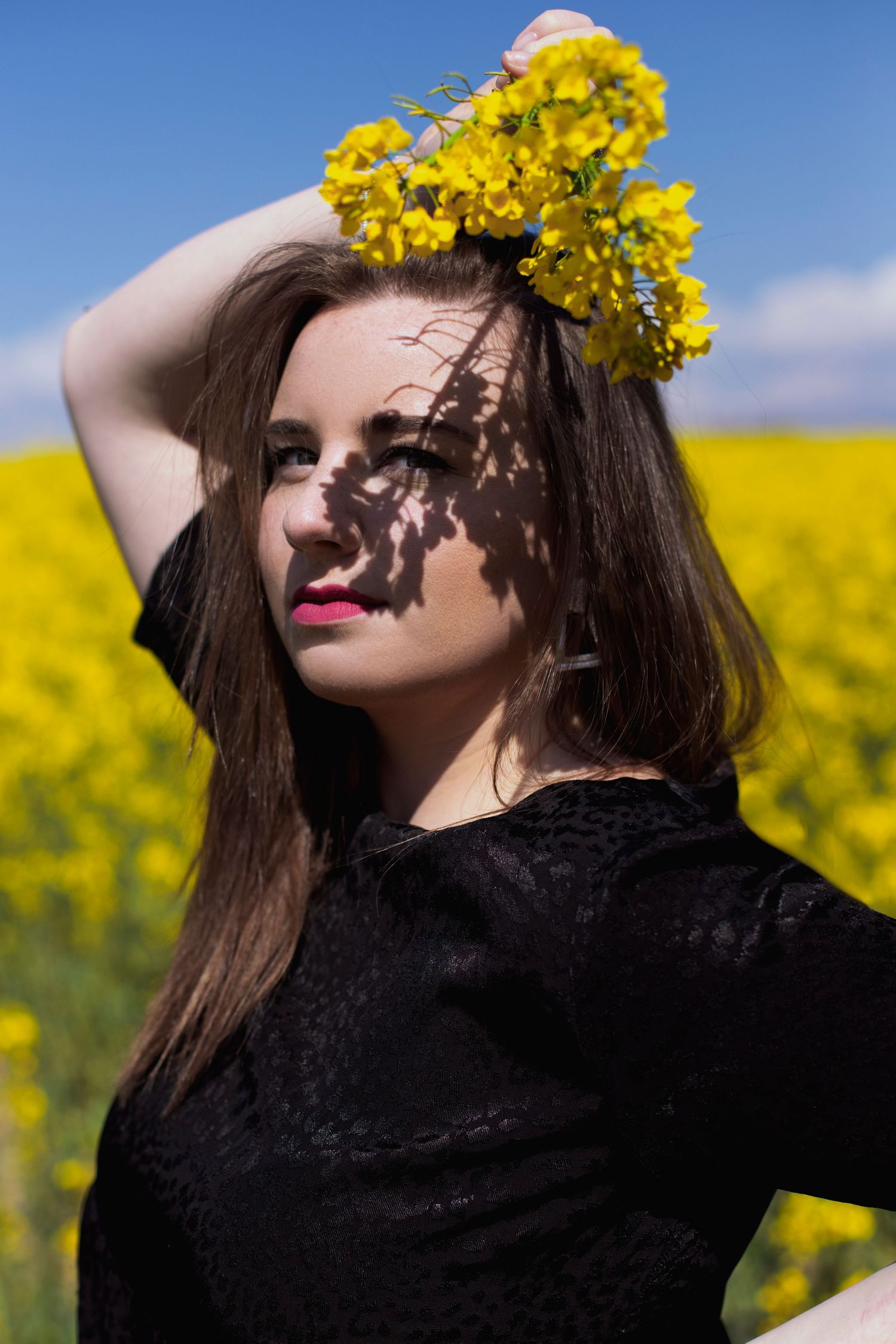 Young woman in black top holding yellow rapeseed flowers as a crown in a sunny field.