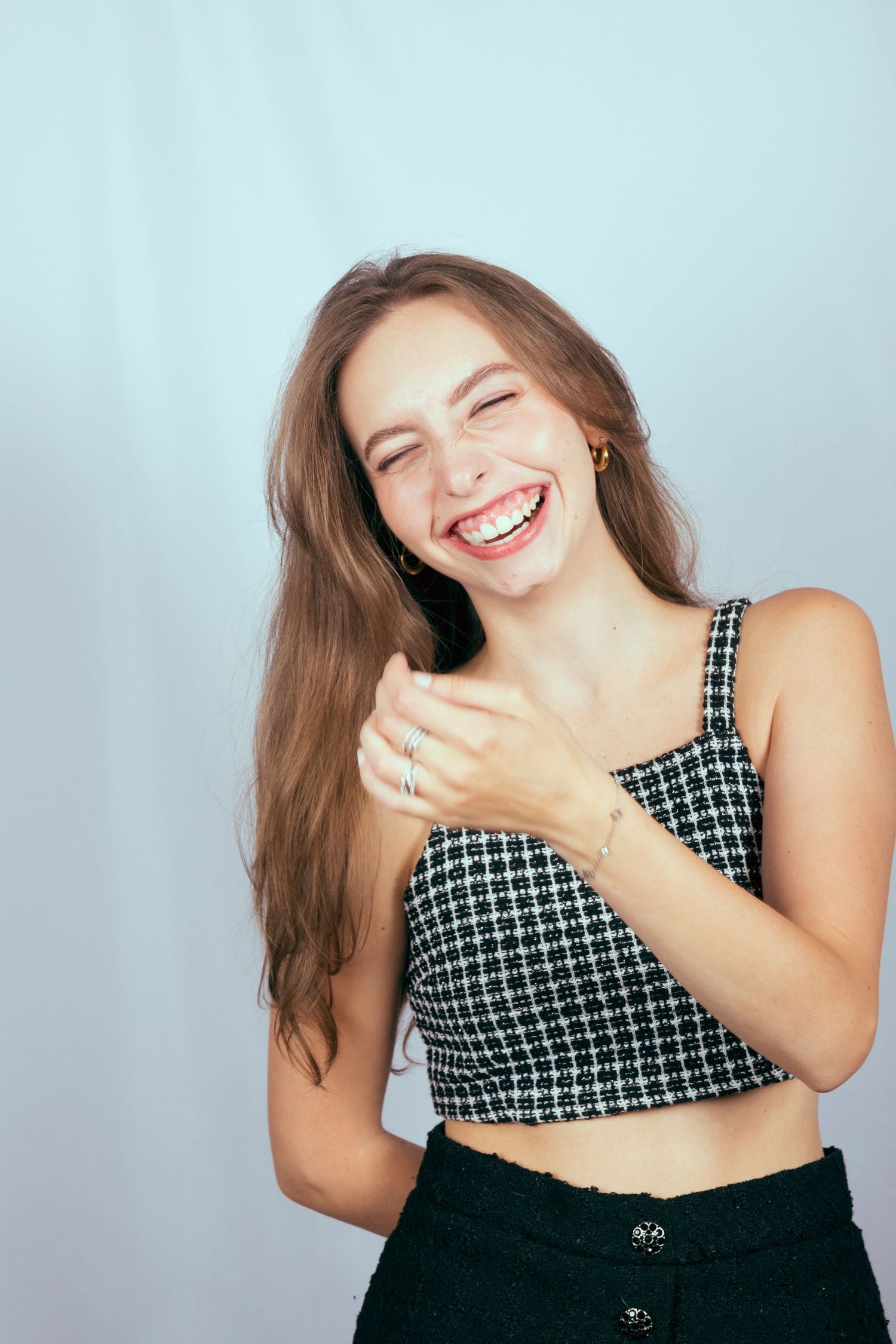 Young woman with long brown hair laughing joyfully, wearing a black and white tweed crop top and black skirt.