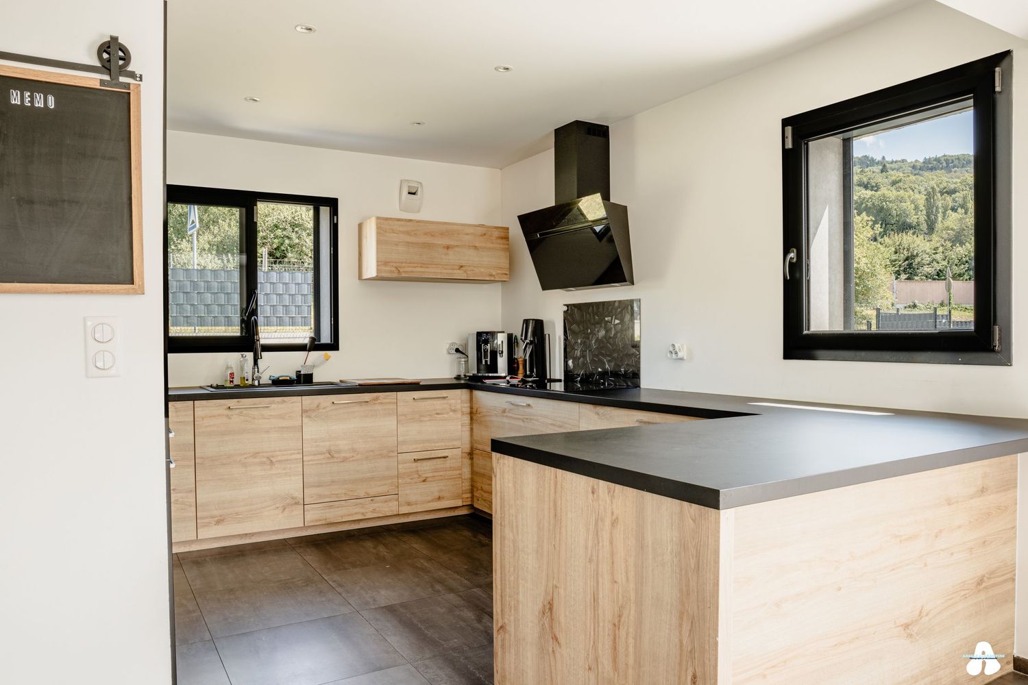 Modern L-shaped kitchen with light wood cabinets, dark countertops and black range hood near windows.
