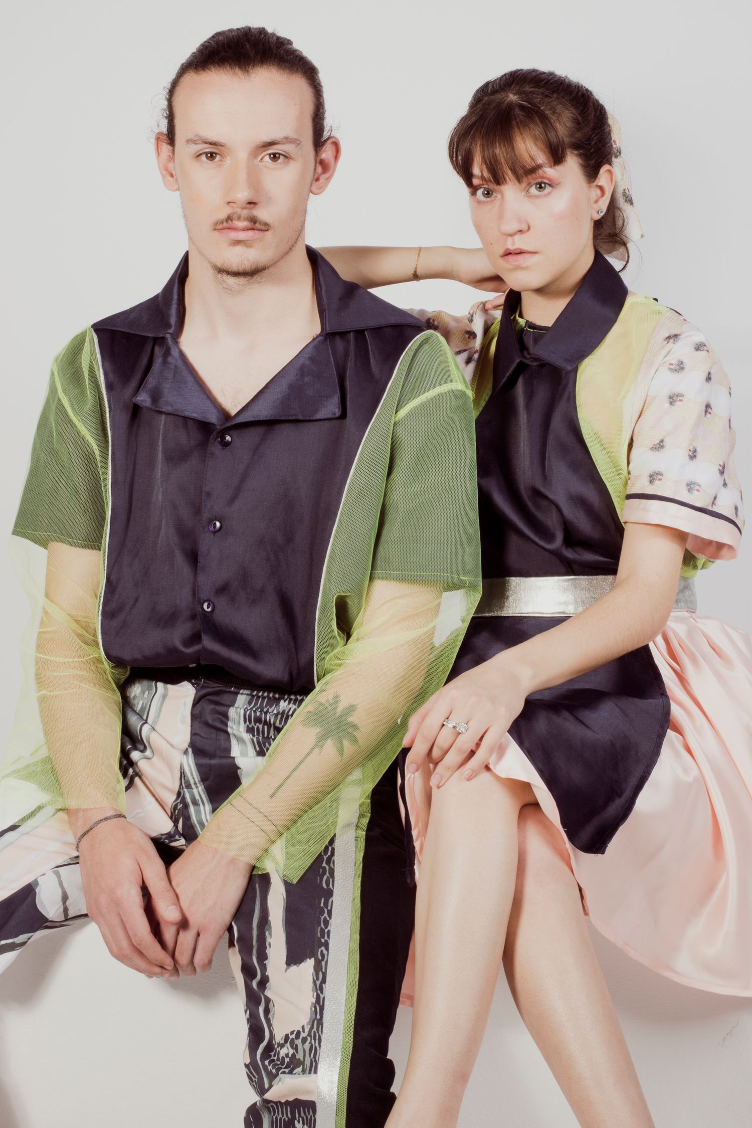 A man and woman model coordinating avant-garde fashion outfits in a white studio setting.