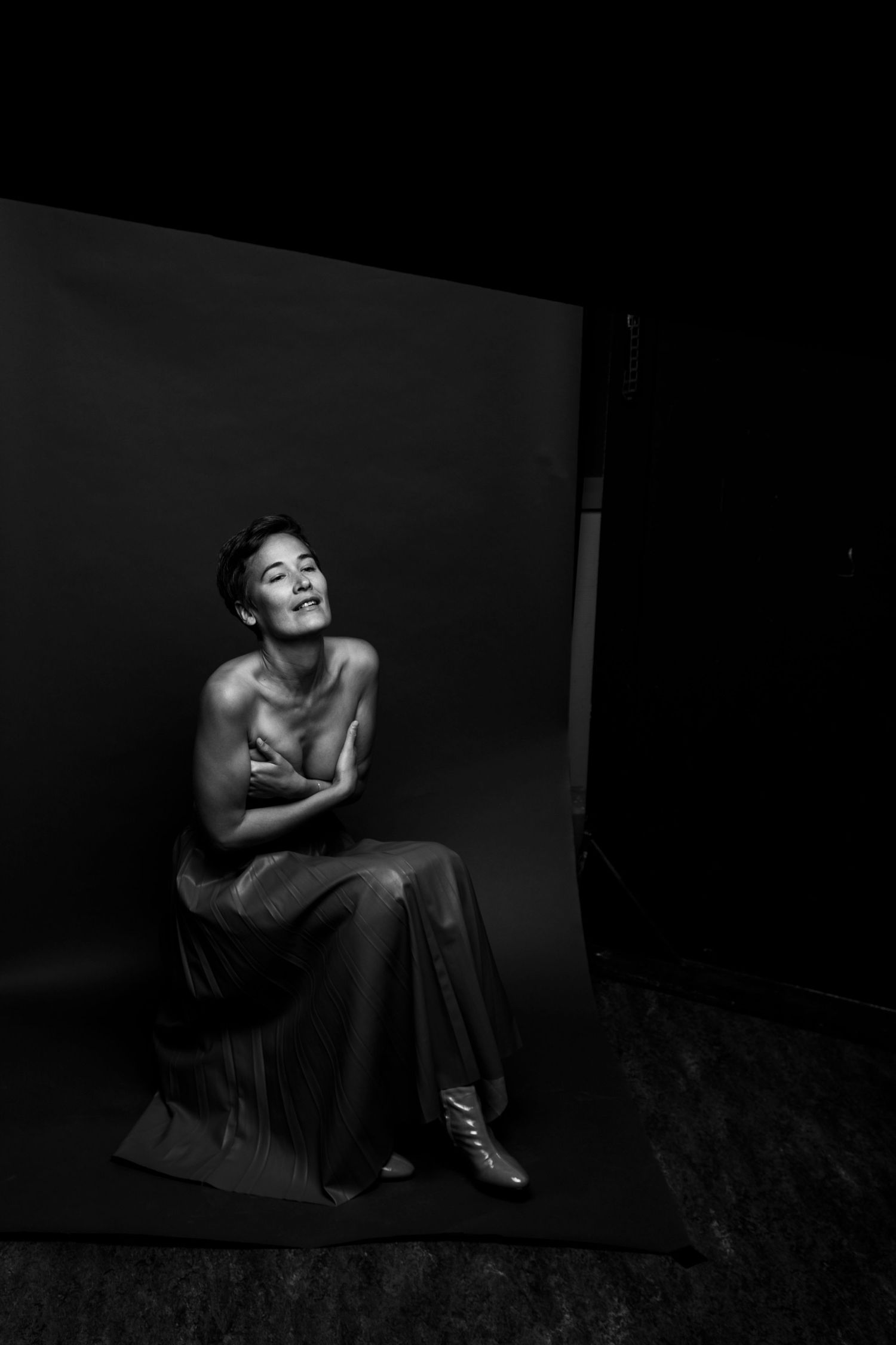 Black and white studio portrait of topless woman seated on stool against dark backdrop.