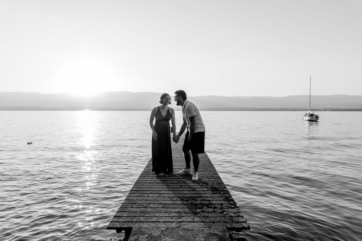 Black and white photo of a couple kissing on a pier at sunset with a sailboat on the lake.