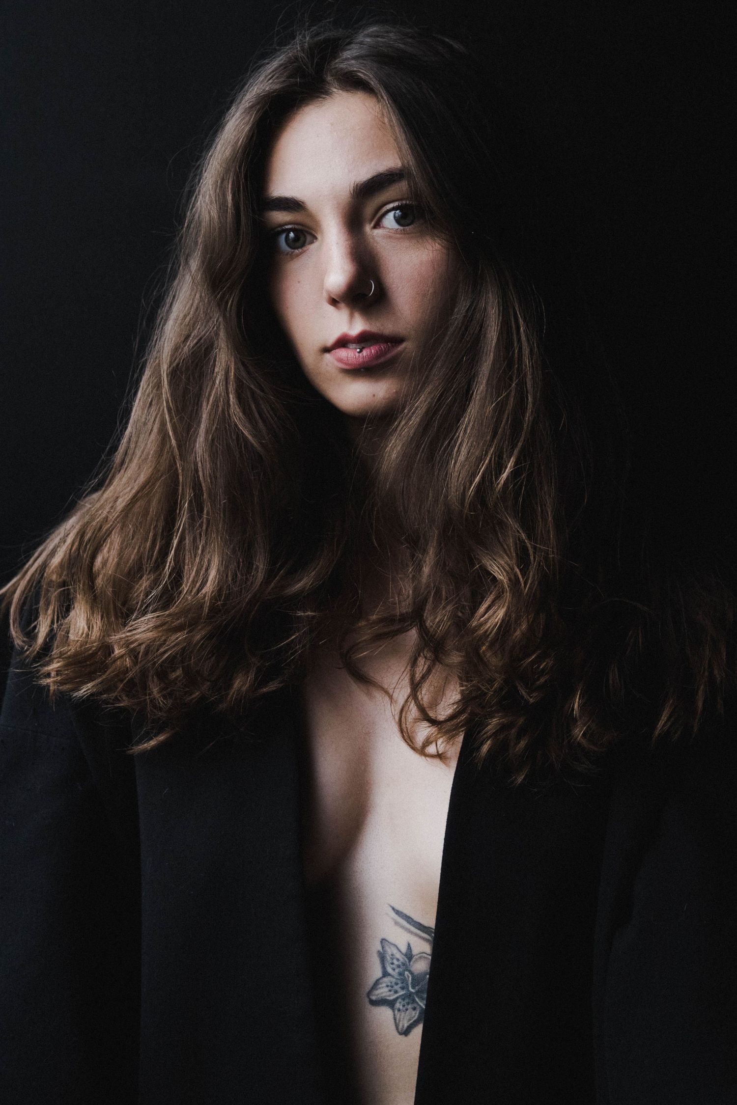 Young woman with long wavy brown hair wearing open black blazer with floral chest tattoo against dark background.