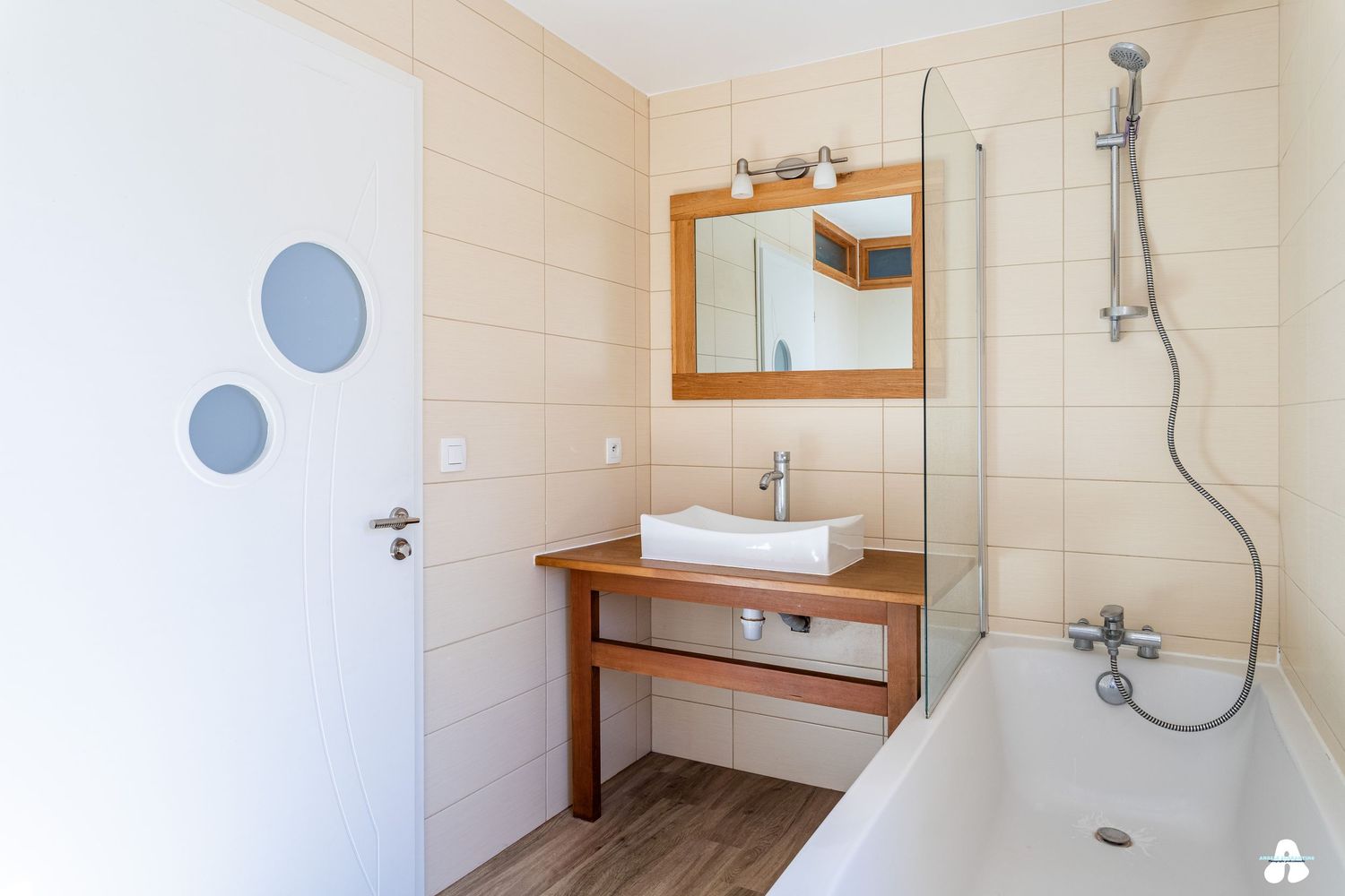 Clean modern bathroom with beige tiles, wooden vanity, wall mirror, walk-in shower and white bathtub.