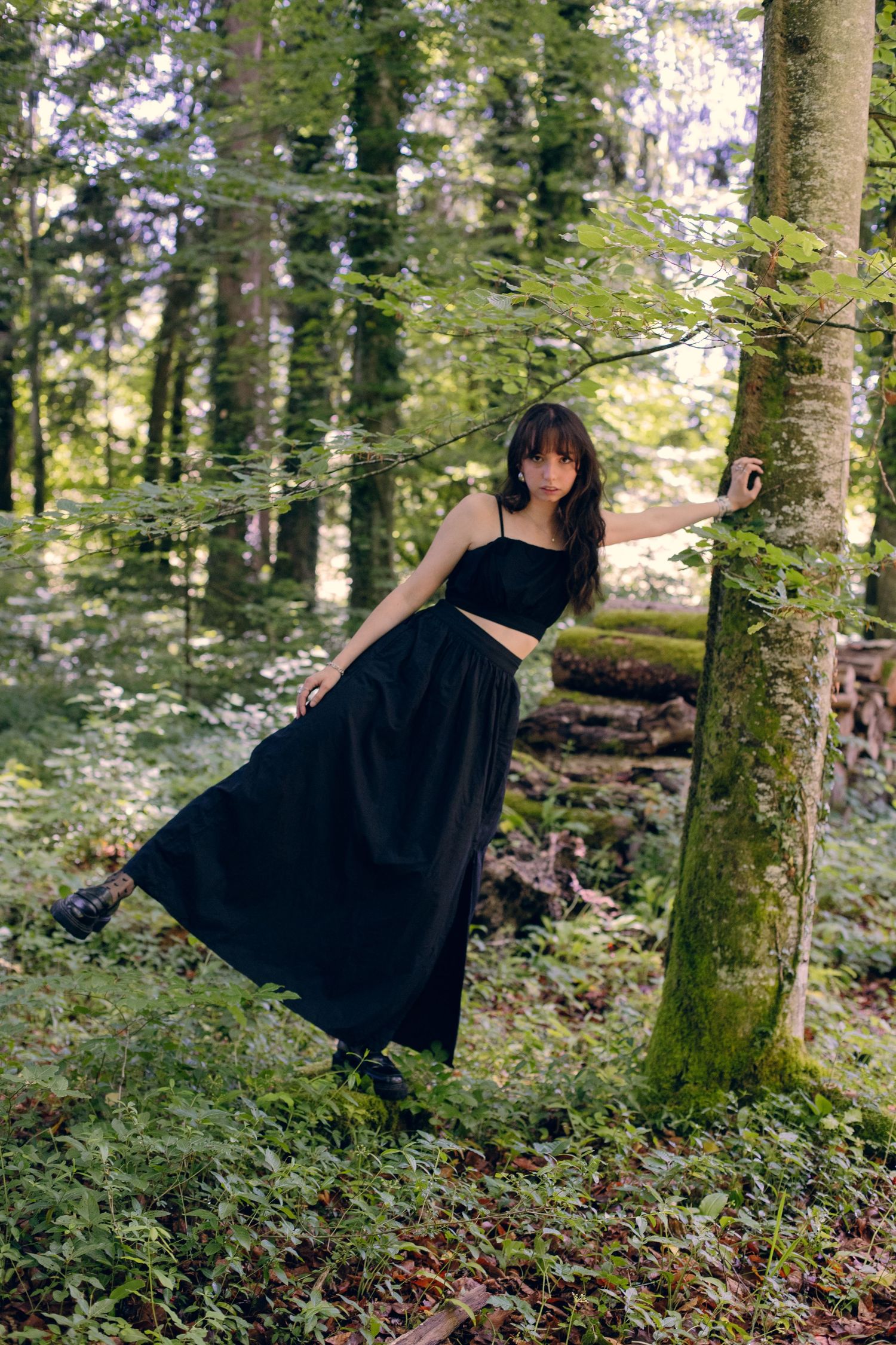 Woman in black outfit leaning against moss-covered tree trunk in lush green forest.