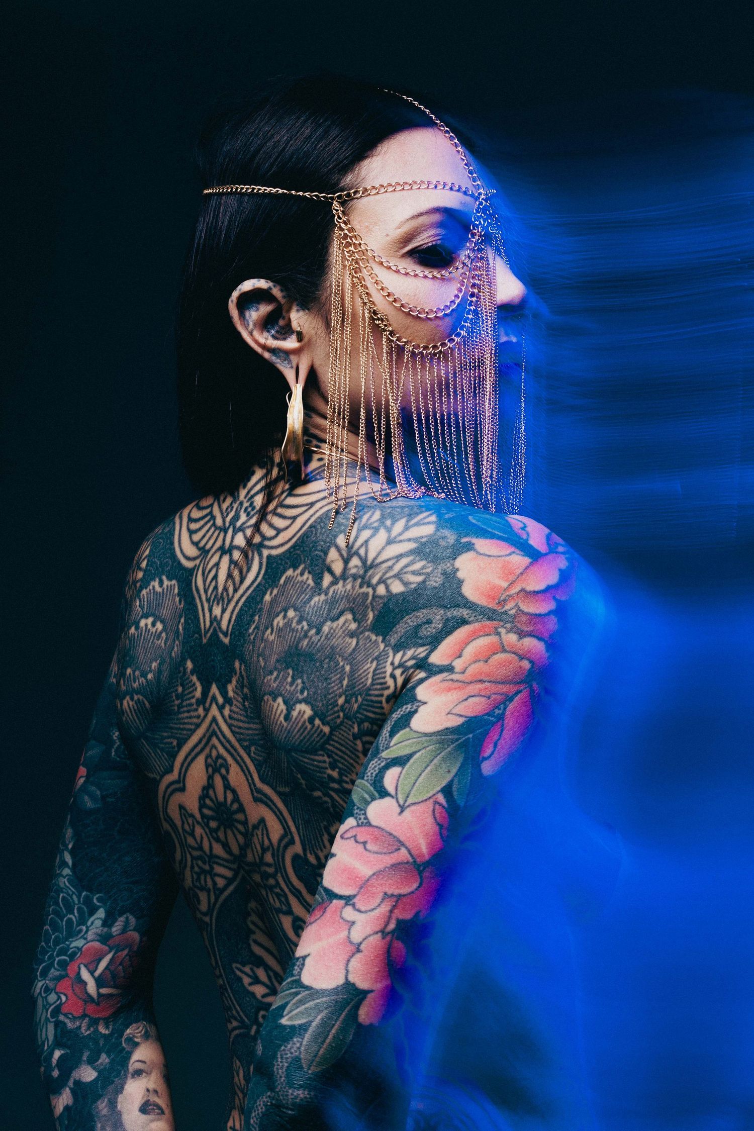 Back view of heavily tattooed woman wearing gold chain headpiece under blue studio lighting.