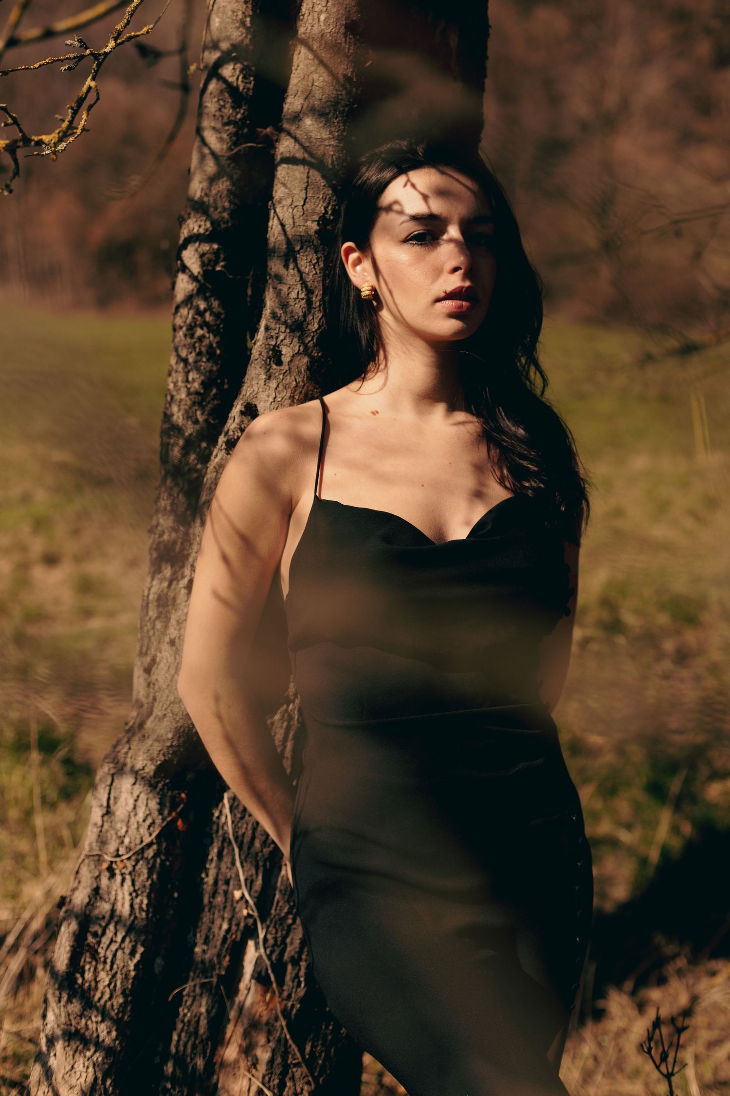 Young woman in black slip dress leaning against tree trunk in natural outdoor setting.