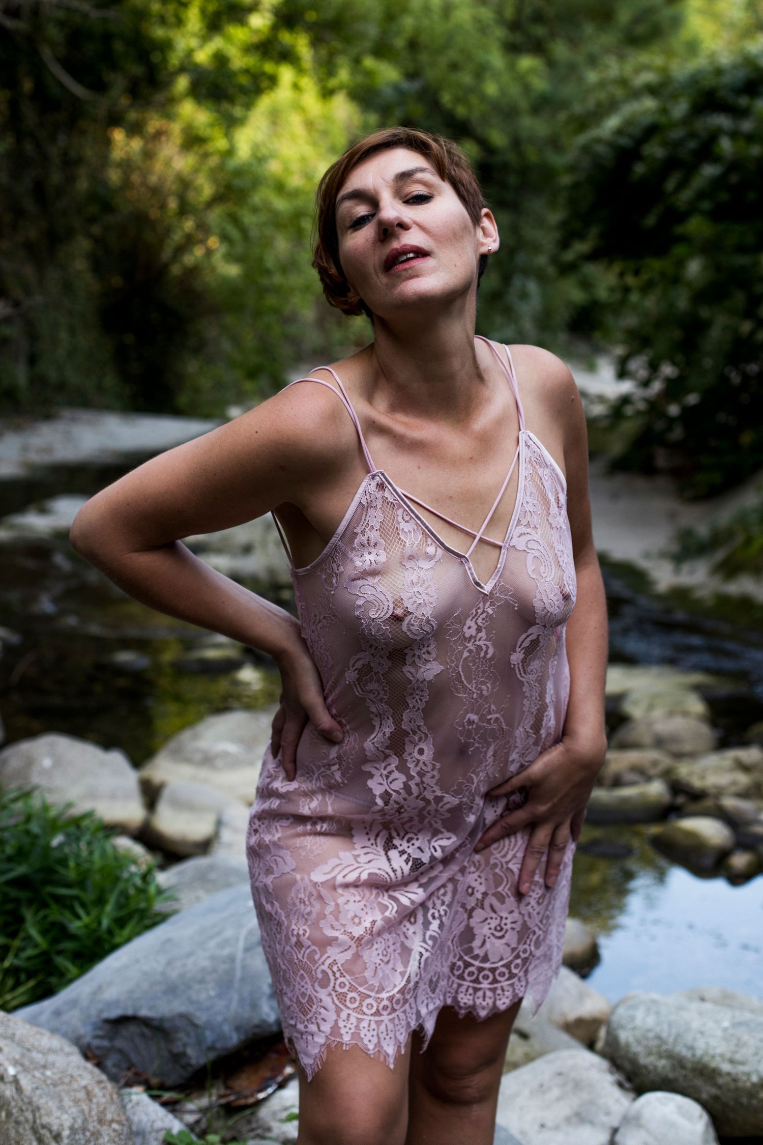 Woman in wet sheer pink lace dress standing in a forest stream.