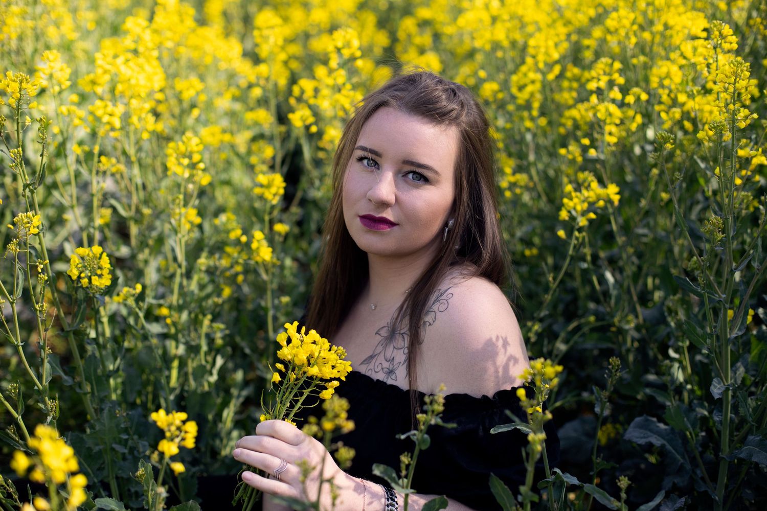 Young woman with tattoos holding yellow flowers posing in a blooming rapeseed field.