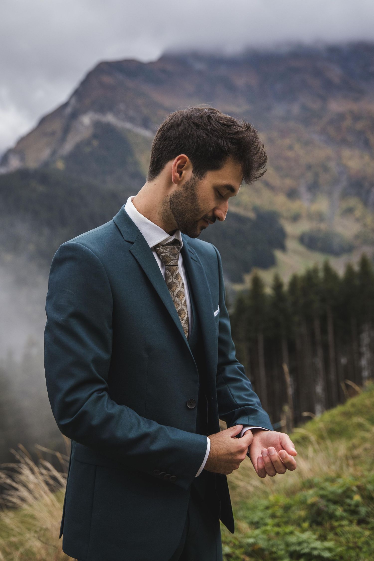 Groom in teal suit adjusting cufflinks with misty mountain backdrop.