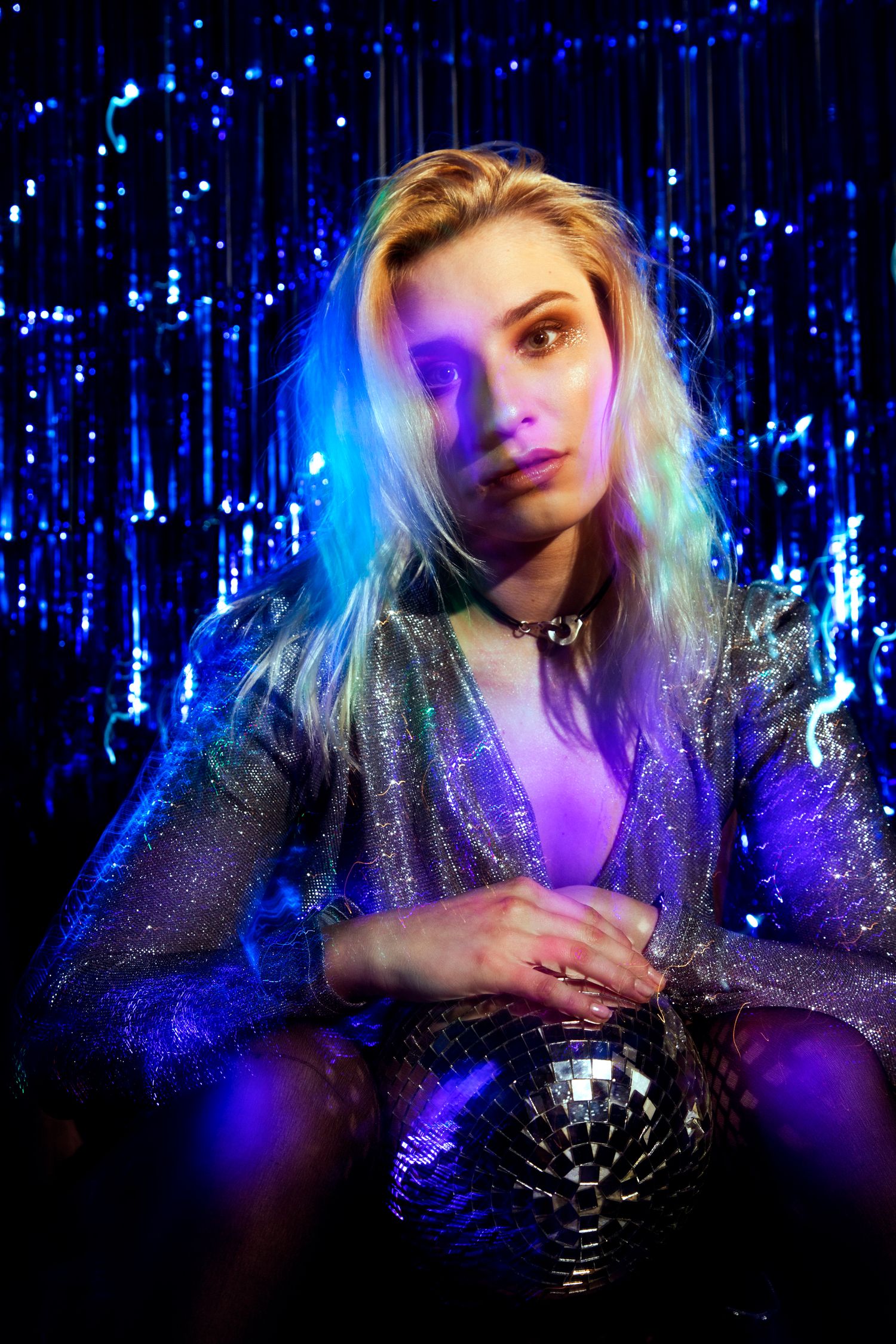 Blonde woman in silver sequin dress holds disco ball under purple and blue neon lighting.