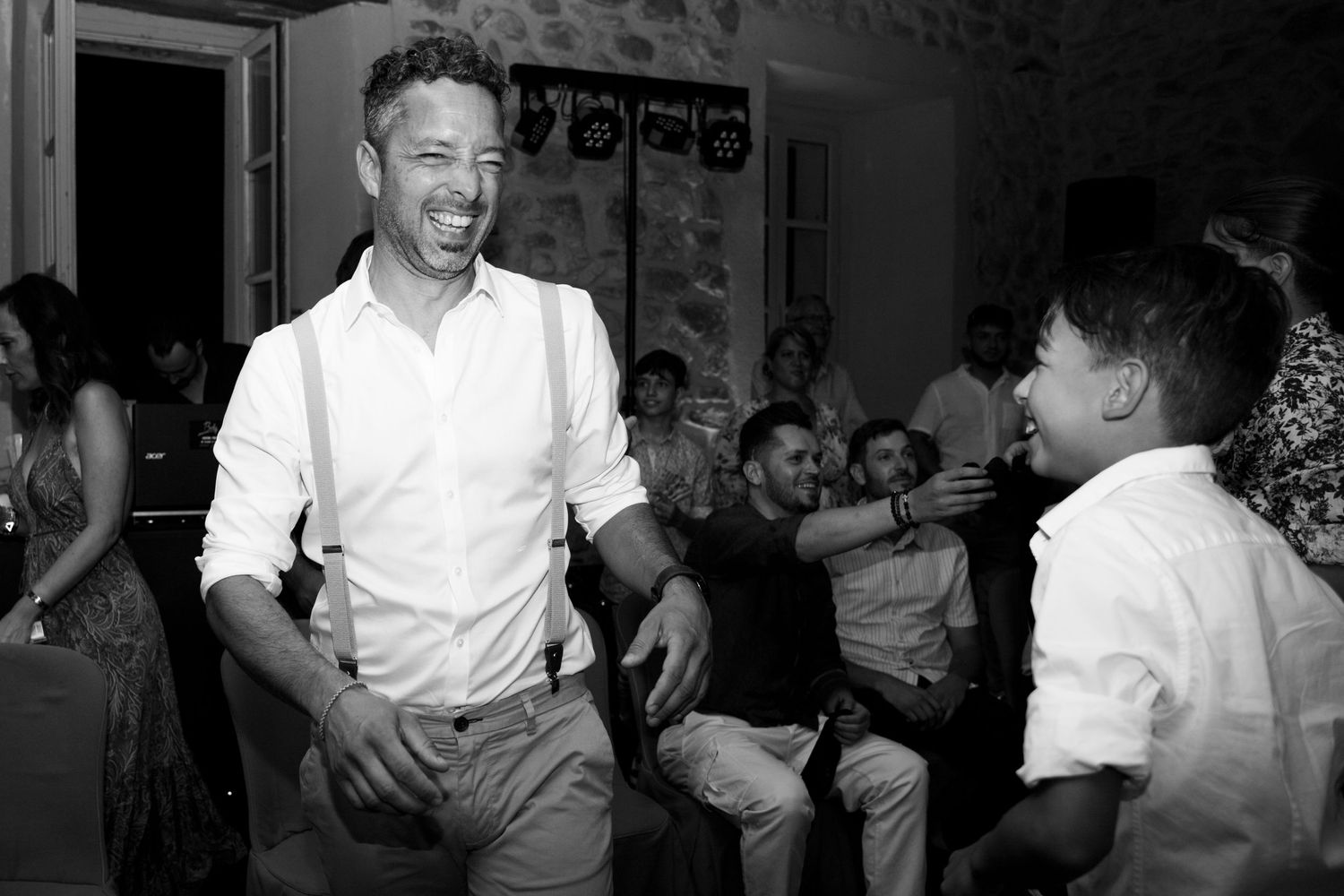 Joyful man in white shirt and suspenders laughing with young boy at wedding reception.