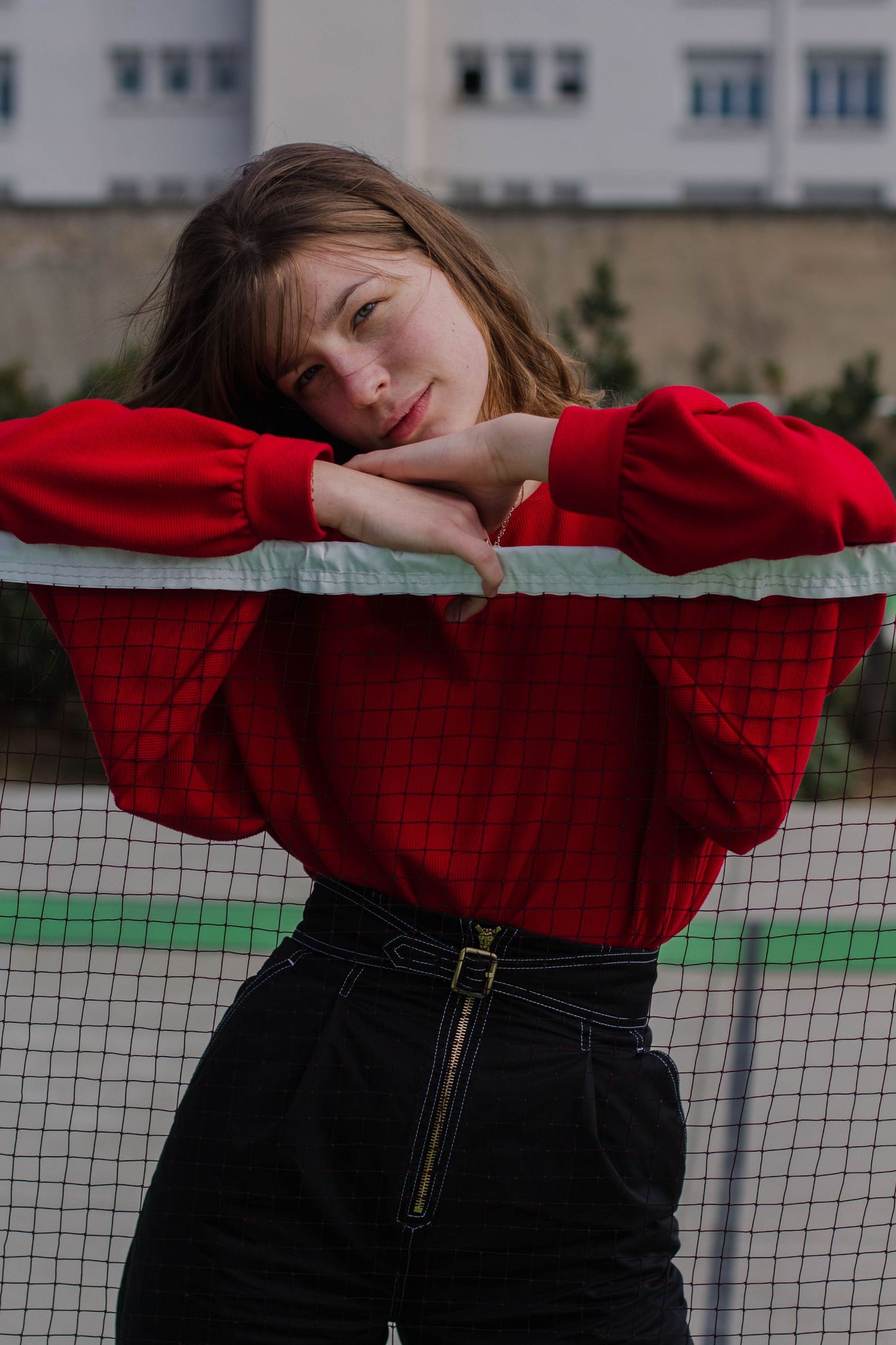 Young woman in red puffed-sleeve top leaning on tennis net at outdoor sports court.